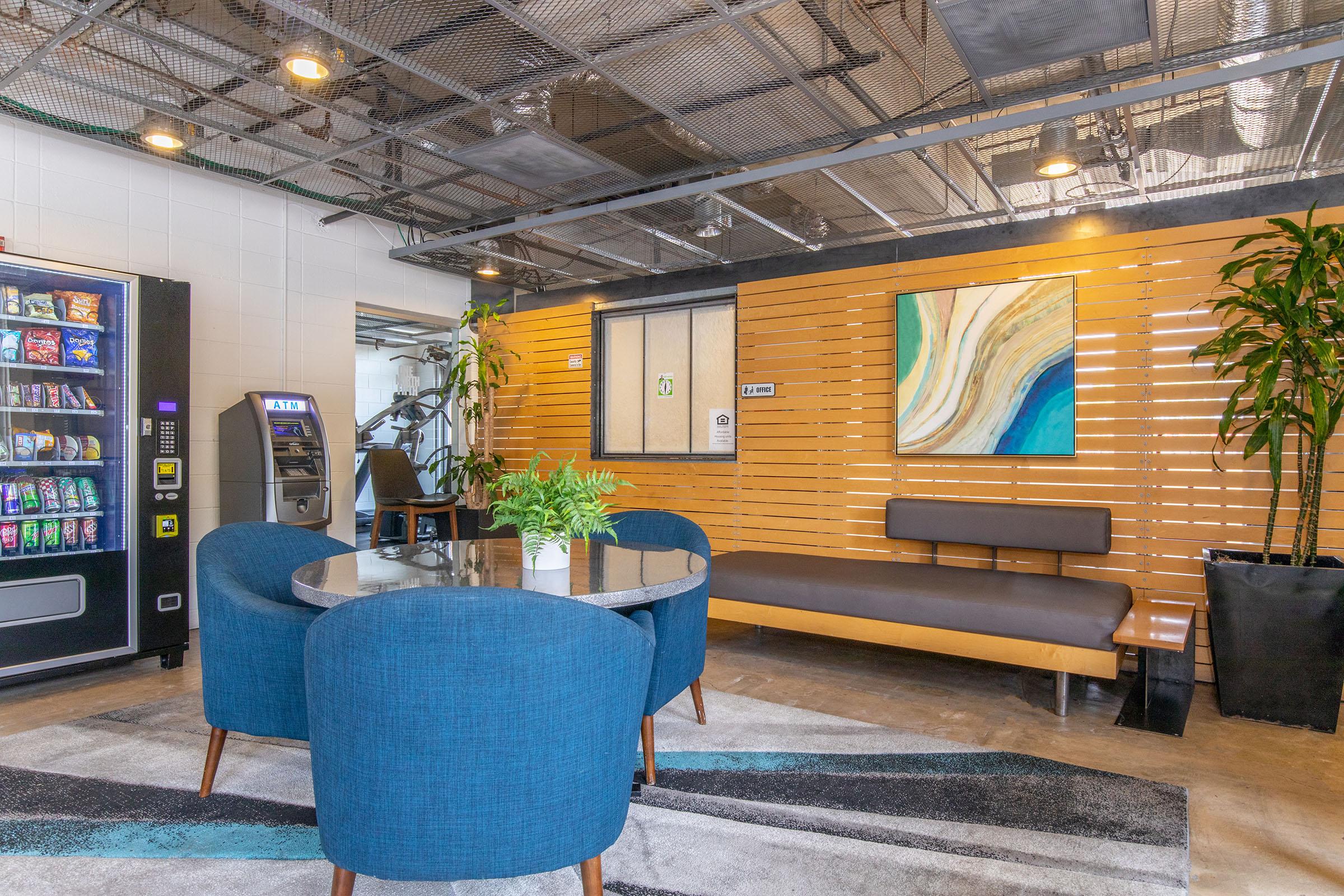 A modern lounge area featuring a round table with three blue upholstered chairs, a long brown bench against a wooden wall, a large abstract painting, a potted plant, and a vending machine. The space has exposed ceilings and a contemporary design, creating a welcoming atmosphere for relaxation or casual meetings.