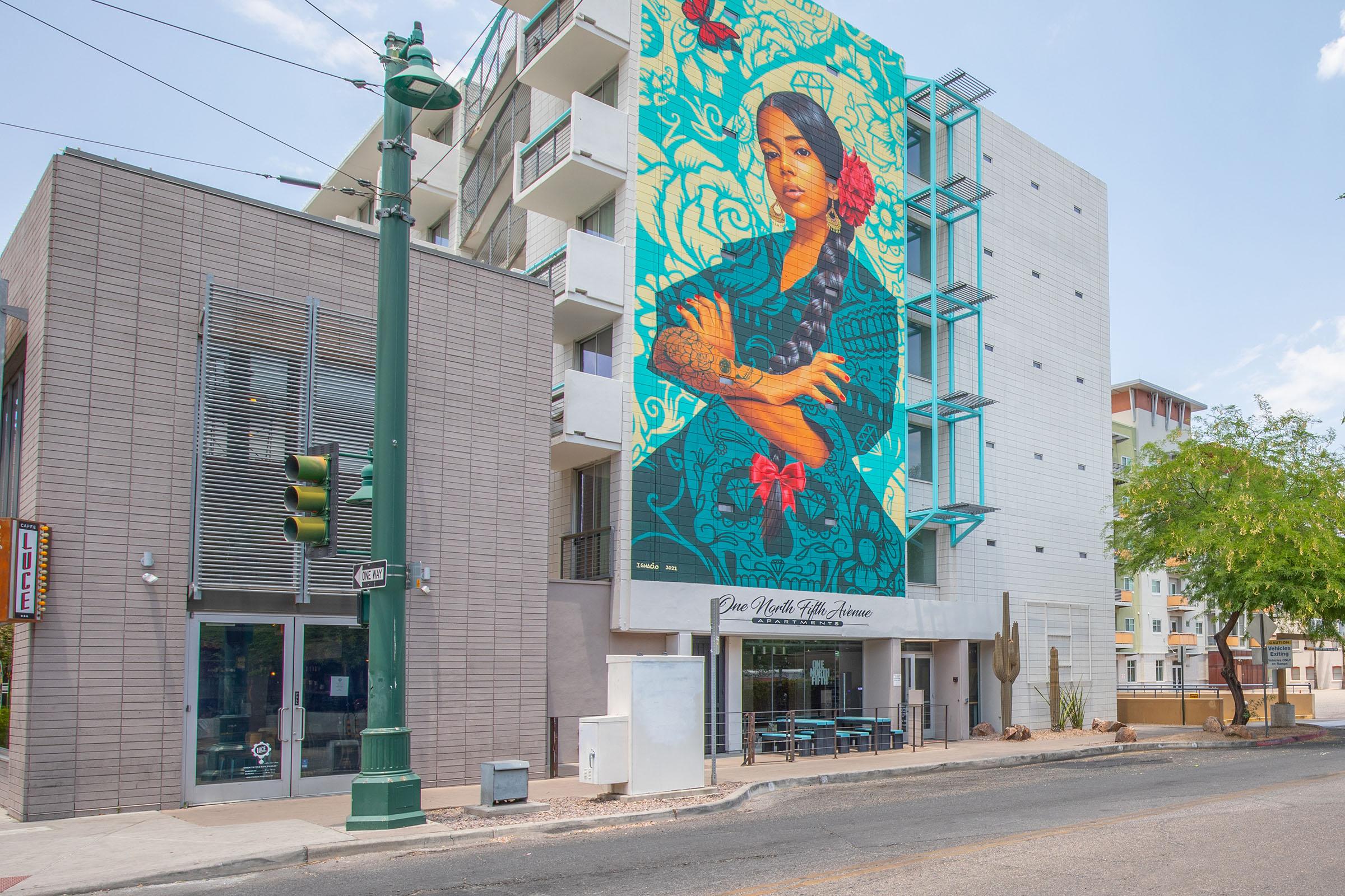 A colorful mural of a woman with a floral design is displayed on the side of a modern building. The street scene features a green traffic light and cacti planted along the sidewalk, with a café visible at the base of the building. The overall atmosphere is vibrant and urban.