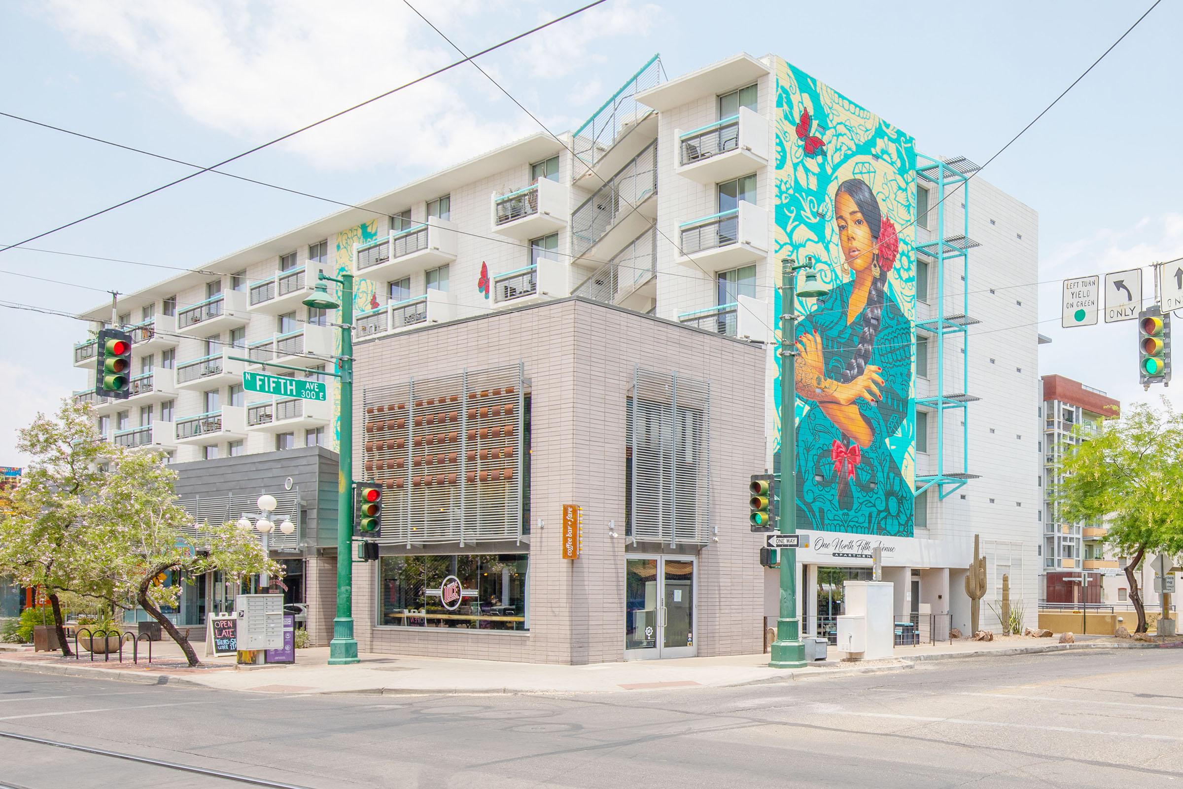 A colorful mural depicting a woman is painted on the side of a modern building with multiple balconies. The image captures a street corner with traffic lights and signage, showcasing urban architecture and art in a vibrant city setting. A tree adds greenery to the foreground.