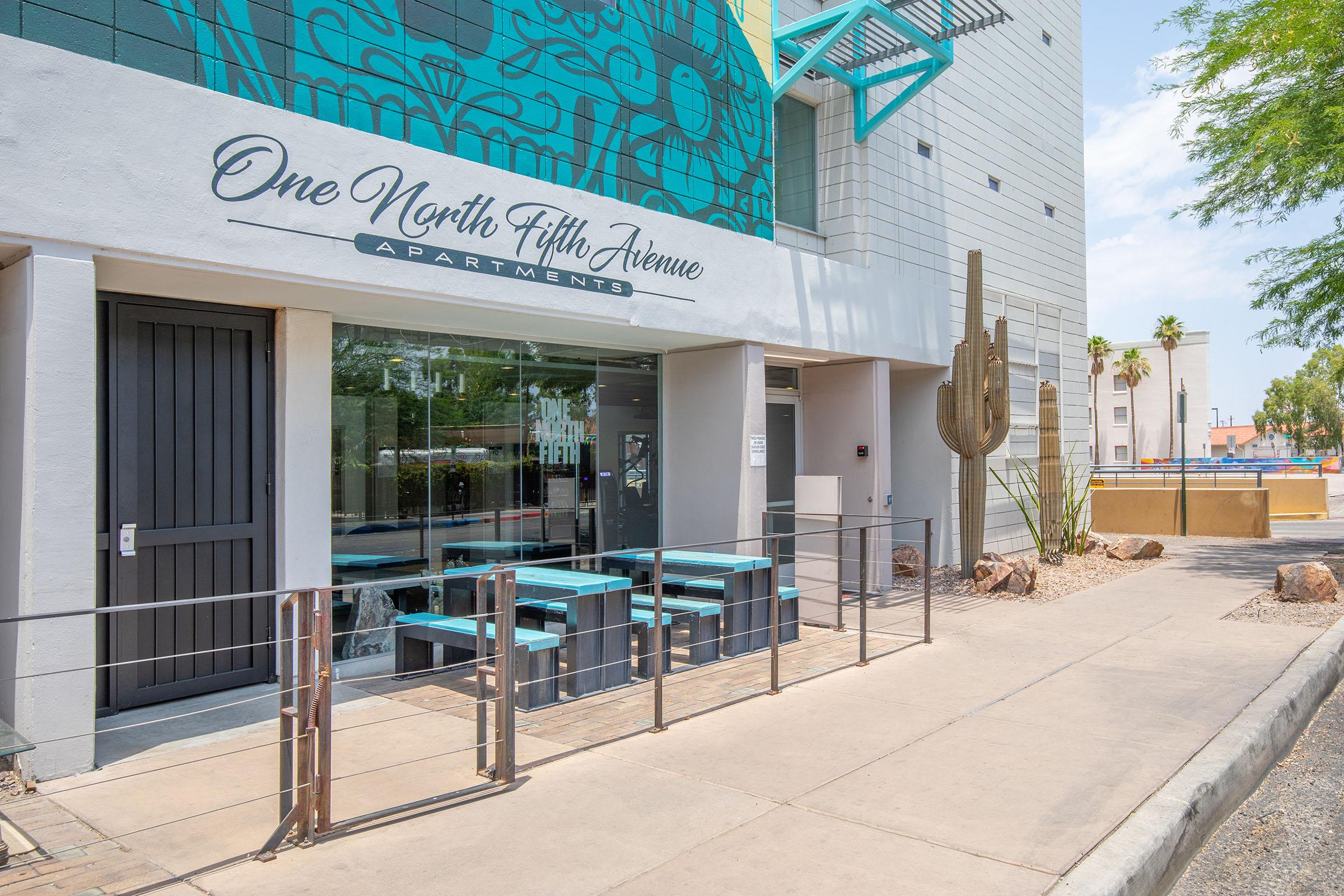 Exterior view of One North Fifth Avenue Apartments, featuring a modern entrance with large glass windows, a decorated wall, and outdoor seating. Cacti and landscaping are visible along the sidewalk, set in a sunny urban environment.