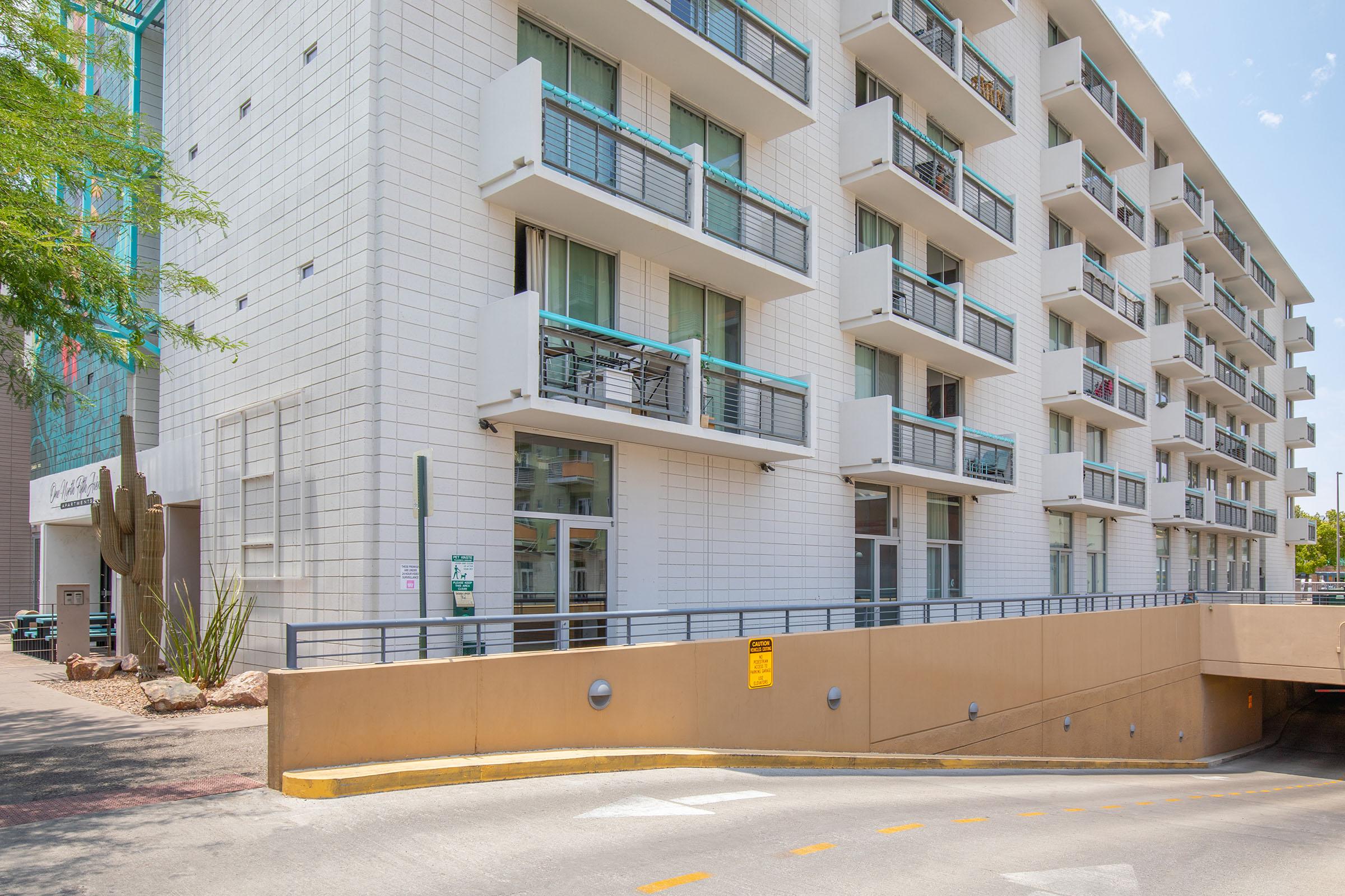 Modern multi-story building with a white exterior, featuring numerous balconies. A ramp leads to an underground parking area, with desert landscaping including a cactus nearby. Clear blue sky above and a few clouds, indicating a sunny day.