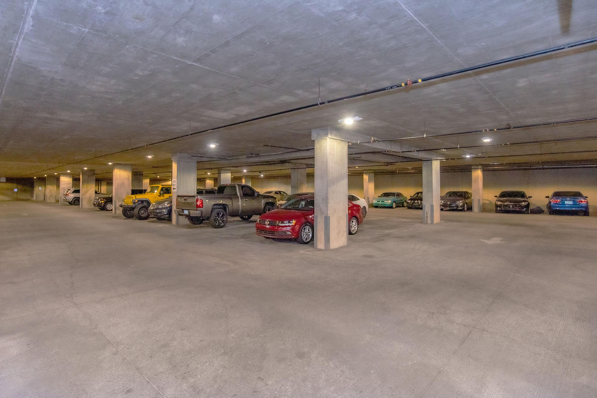 An underground parking garage with several parked cars, including a red sedan, a black truck, and various other vehicles. The concrete structure features bright overhead lighting and wide aisles, creating a spacious and organized environment.
