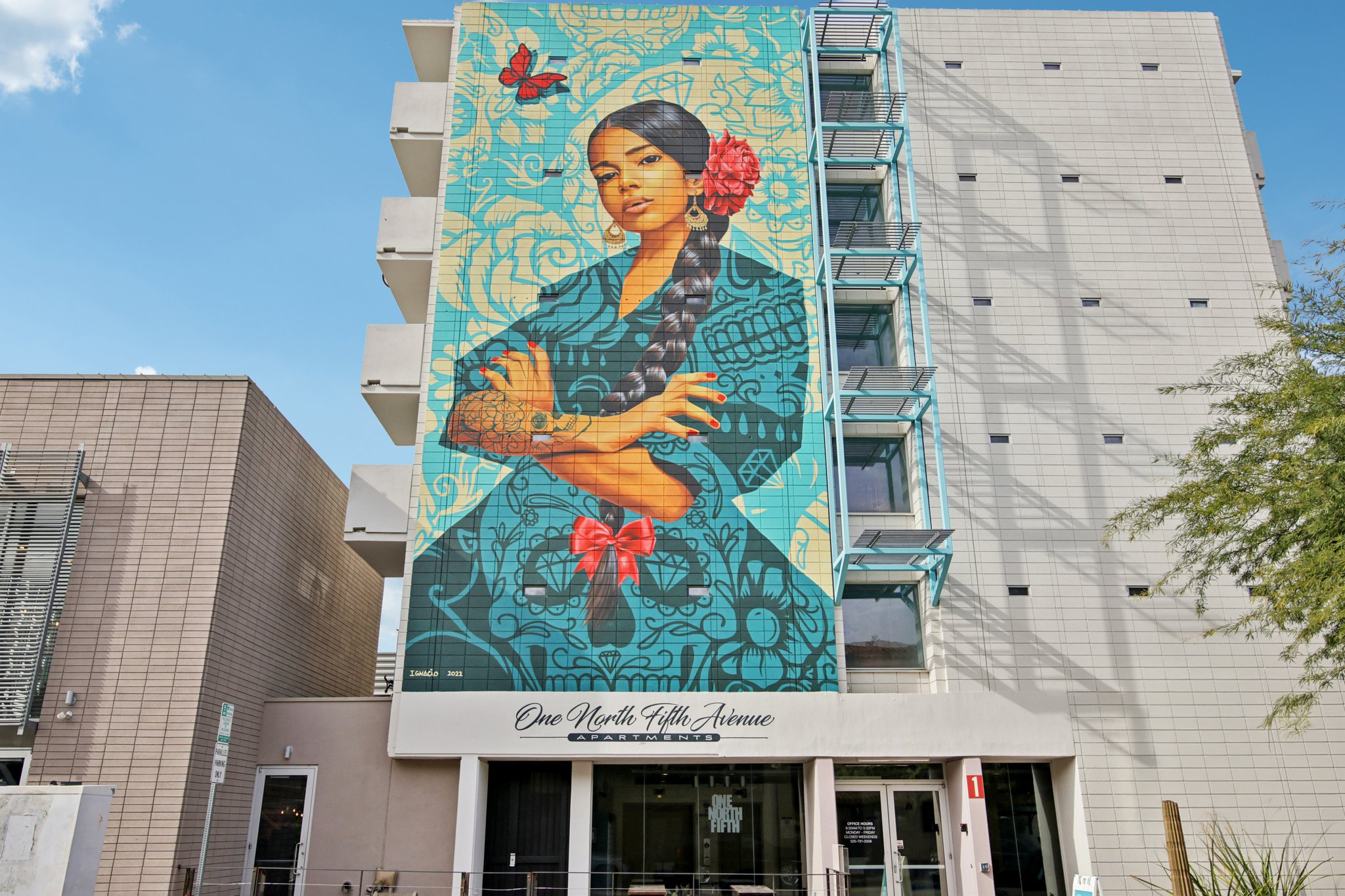 A colorful mural of a woman with long, braided hair and tattoos adorns the side of a modern building. She is depicted with a bold expression, surrounded by floral patterns and a butterfly. Below the mural, the building's entrance is visible, labeled "One North Fifth Avenue." The sky is clear and blue.