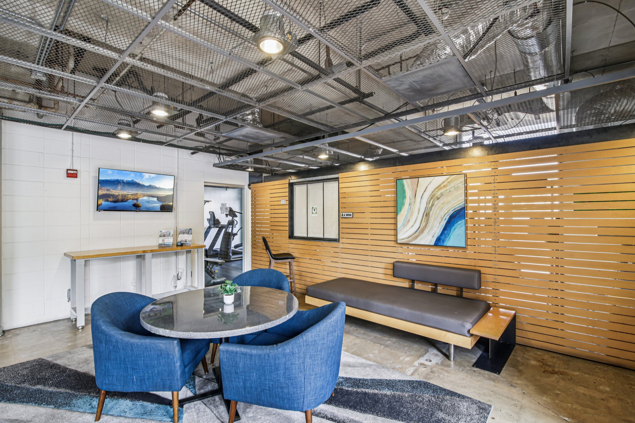 A modern lounge area featuring a round table with four blue chairs, a sleek black bench, and wooden wall panels. A small potted plant sits on the table. In the background, there is a fitness area visible with gym equipment and a framed landscape art piece on the wall.