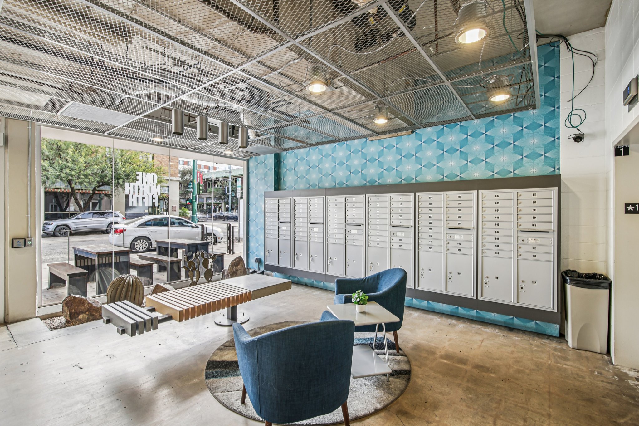 Modern lobby interior with blue geometric wall design, featuring mailboxes against one wall. There are two blue armchairs and a low wooden table in the center. Natural light enters through large windows, and a street view is visible outside.