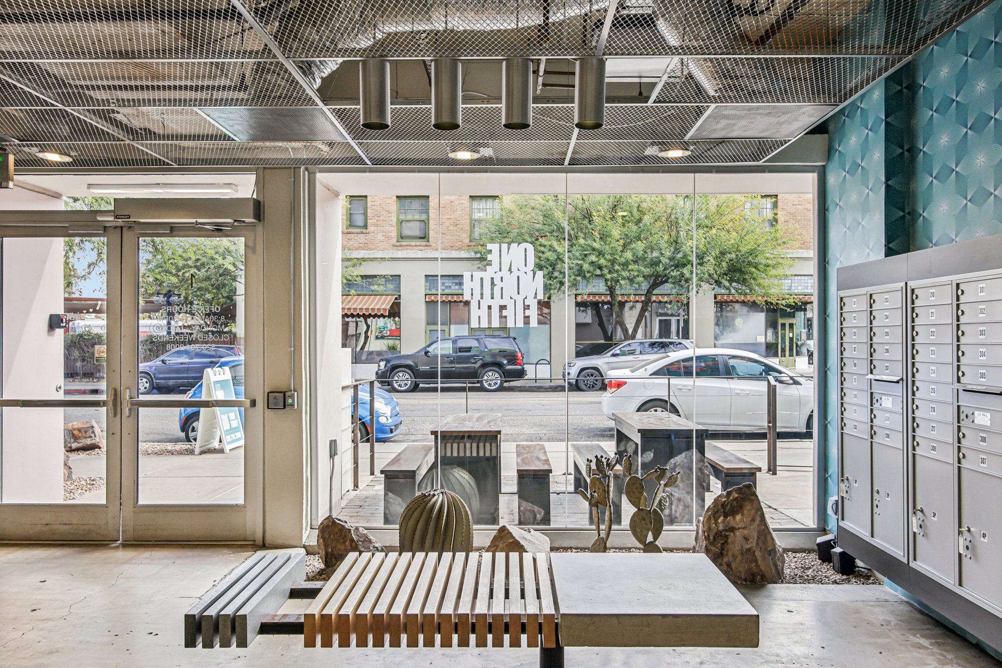 Interior view of a modern lobby with a sleek design, featuring a reception area, mailboxes, and large glass windows showcasing a street scene outside. Decor includes natural elements like plants and stones, creating an inviting atmosphere.