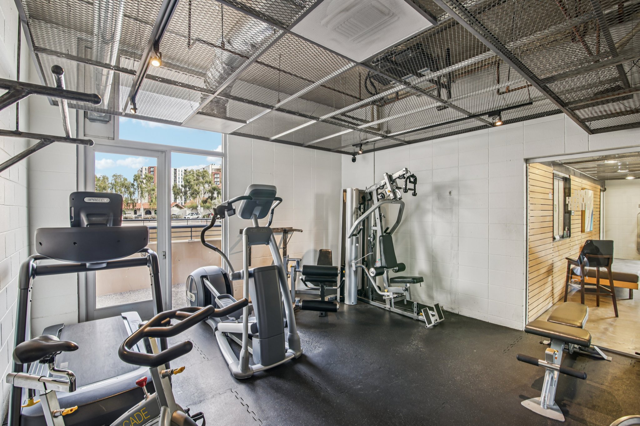 A modern gym space featuring various exercise equipment, including treadmills, stationary bikes, and weight machines. Large windows allow natural light, creating an inviting atmosphere. The floor is covered with black mats, and there are walls with mirrors and wooden paneling.