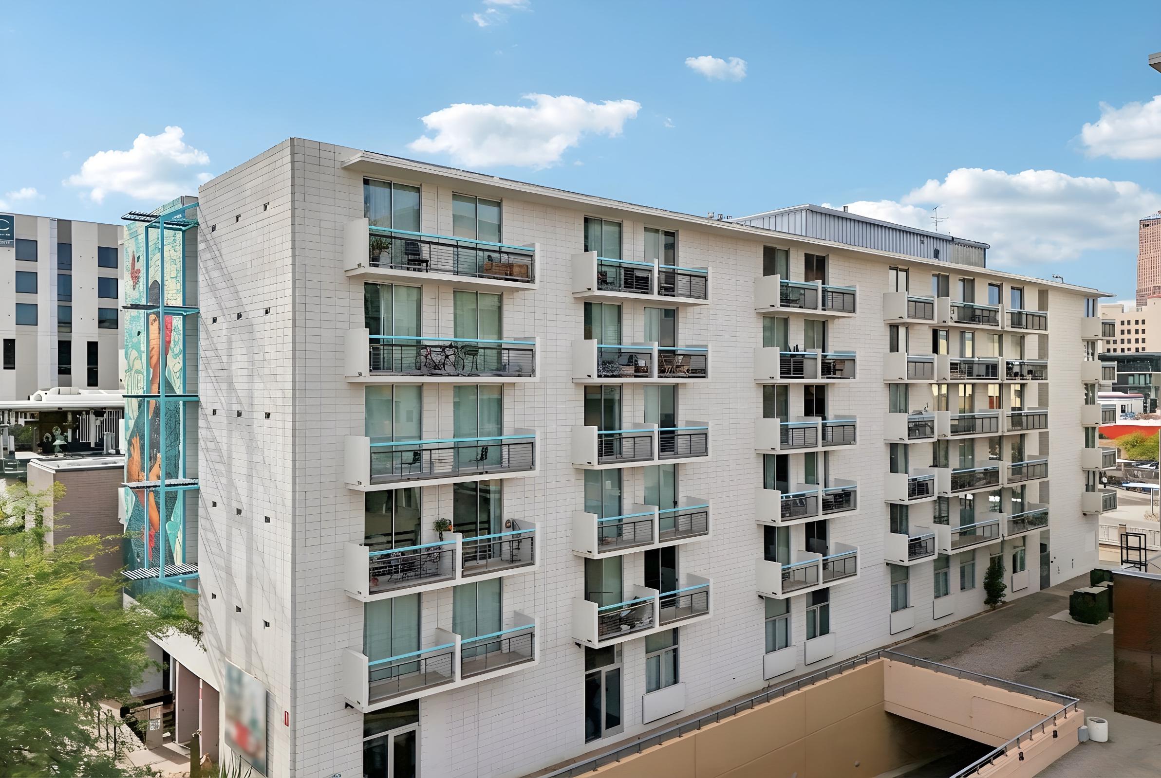 A modern, multi-story apartment building with numerous balconies. The facade is primarily white, and there is colorful artwork displayed on a glass wall at the side. The sky is clear with a few clouds, and the surrounding area shows other buildings and greenery.