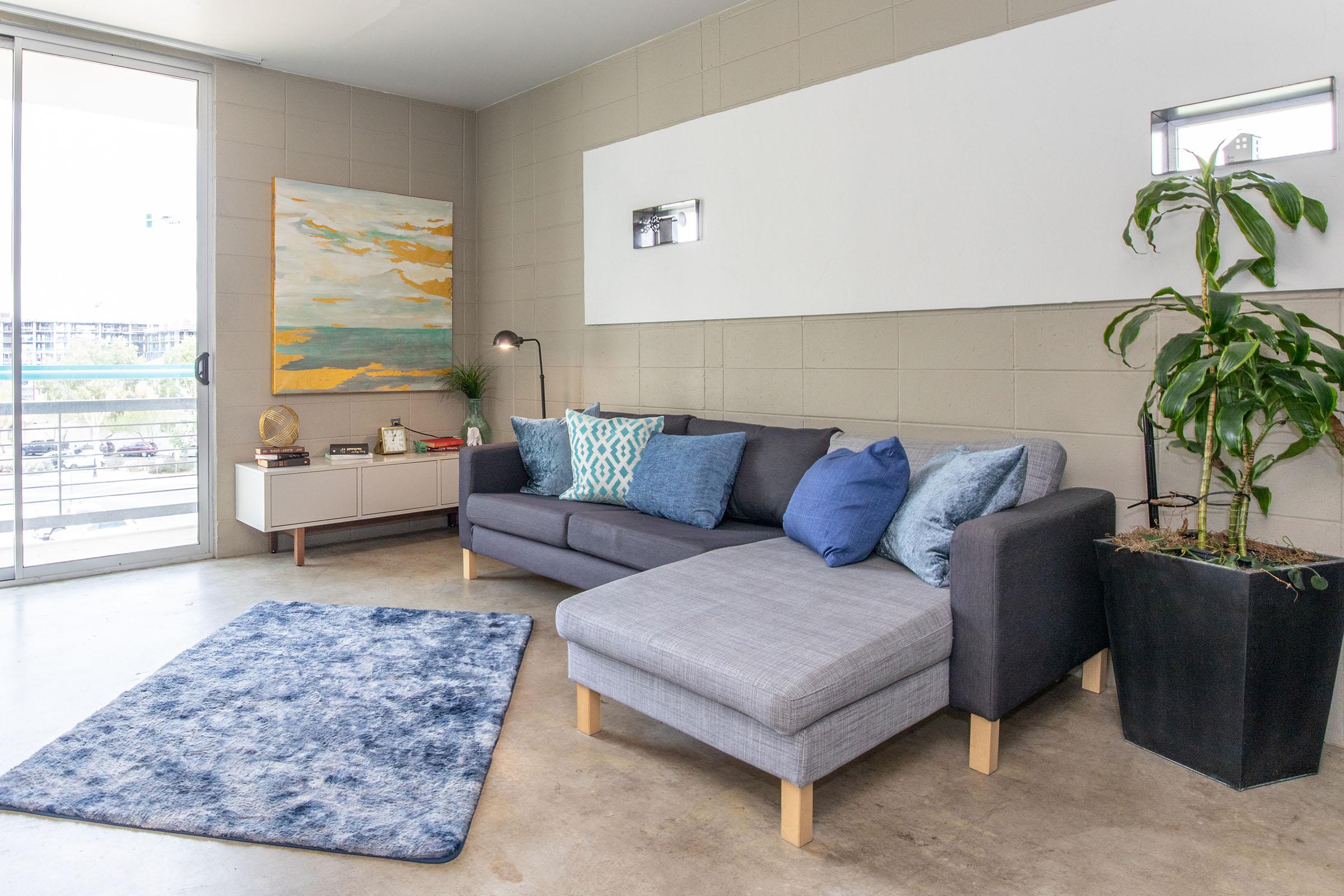 A modern living room featuring a gray sectional sofa with blue accent pillows, a light blue area rug, and a sleek white console table. There's a decorative piece on the wall, a potted plant in a dark planter, and large windows allowing natural light to fill the space.