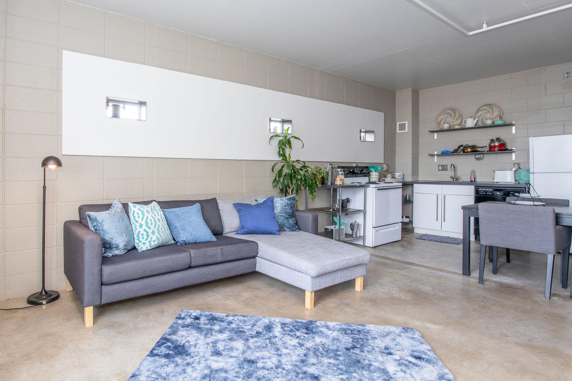 A modern living space featuring a gray sectional sofa adorned with blue and teal cushions, a light gray rug, and a small dining area with a table and chairs. The decor includes a green plant and contemporary kitchen elements against a neutral wall. Illuminated by a standing lamp, the space is bright and inviting.
