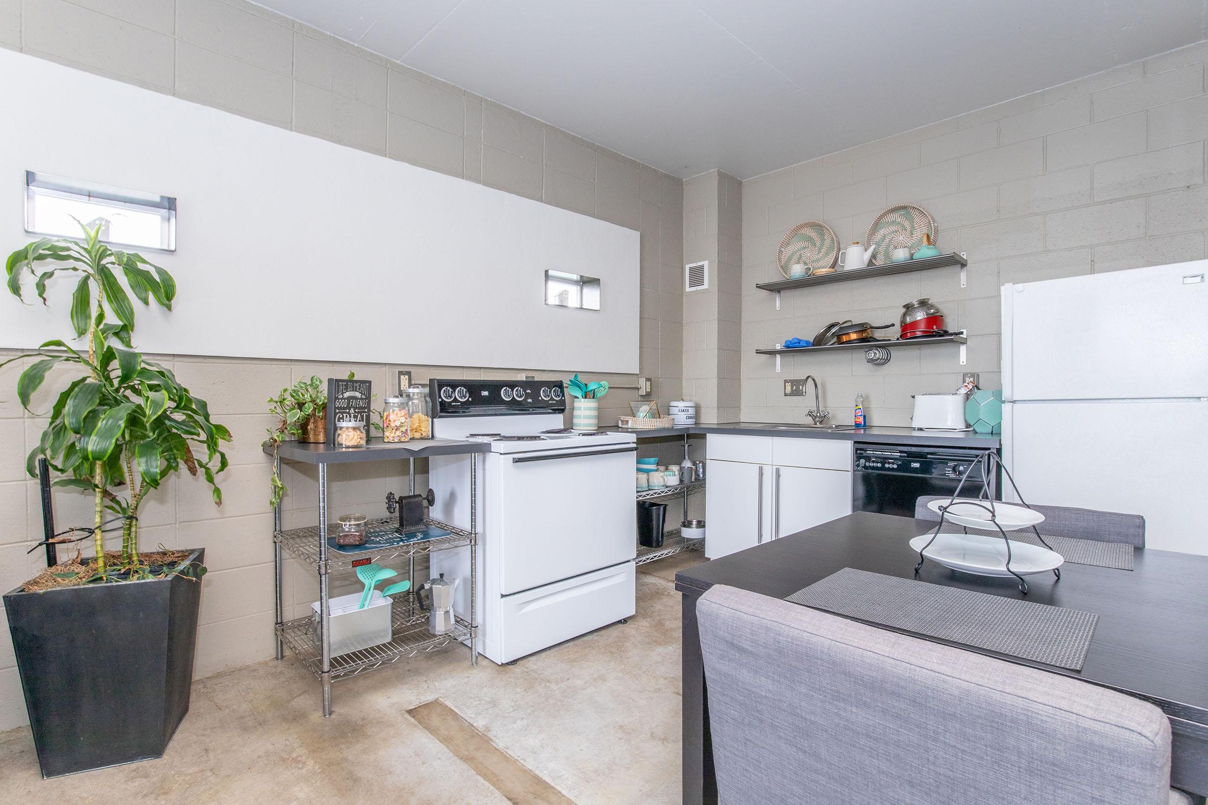 A modern kitchen showcasing a clean and minimalist design. Features include a white refrigerator, a silver stove, and a countertop with cooking utensils. A potted plant adds greenery, while shelves display various kitchen items. A dining table with placemats completes the compact, functional space.