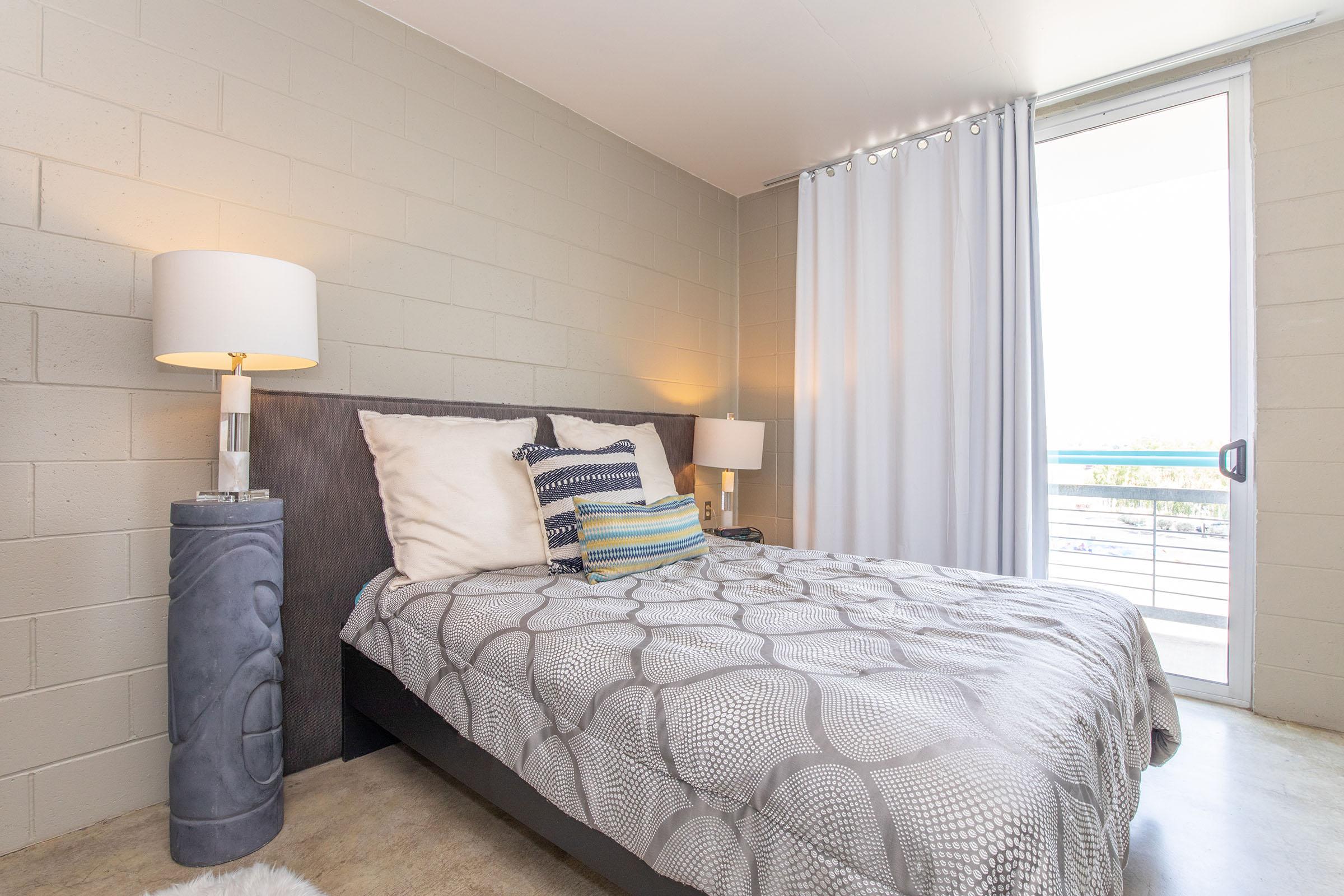 A modern bedroom featuring a neatly made bed with patterned bedding, two bedside lamps, and decorative pillows. Light-colored curtains cover a window that opens to a balcony, while gray walls provide a minimalist backdrop. The floor is smooth, complementing the contemporary aesthetics of the room.