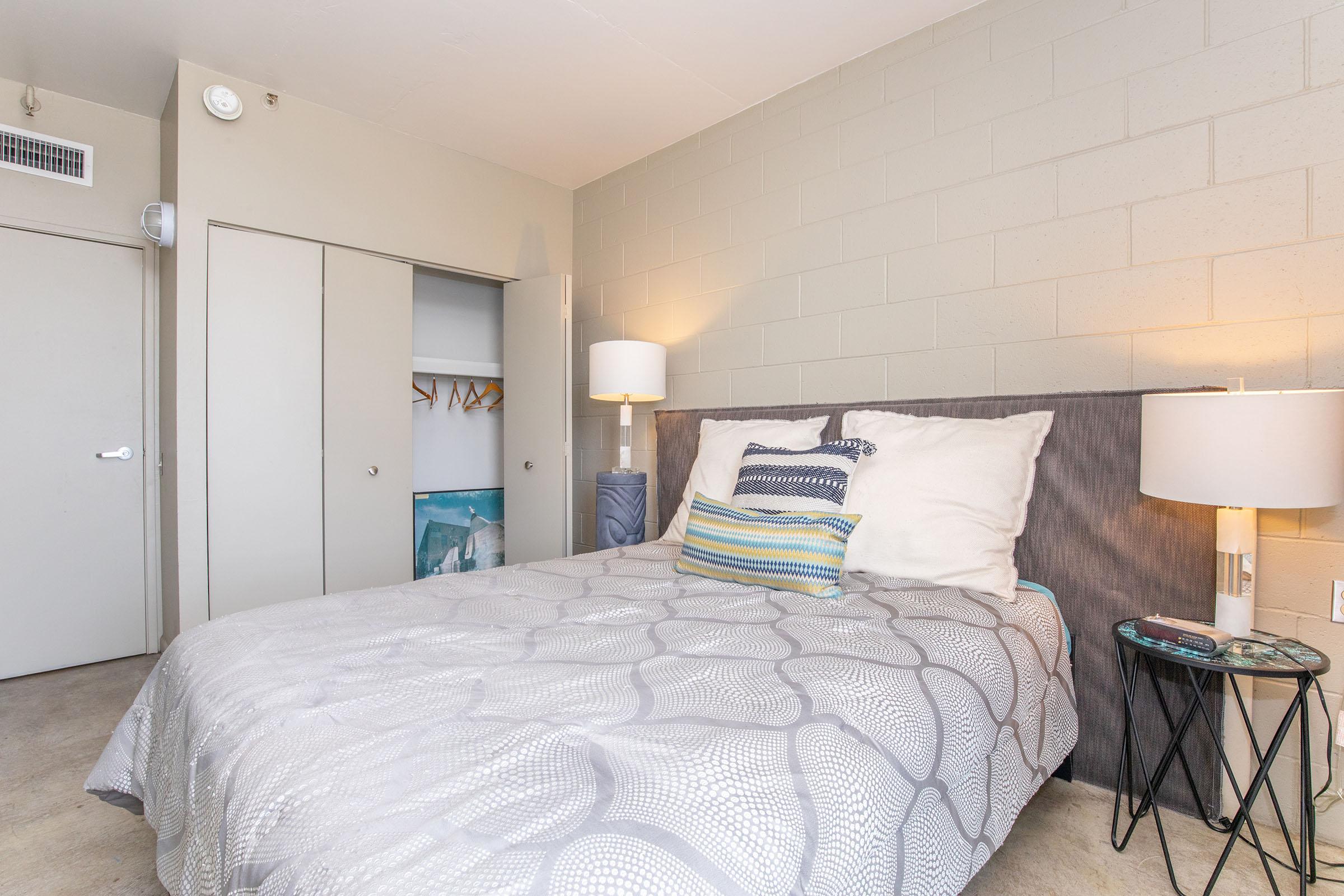 A cozy bedroom featuring a queen-sized bed with a gray patterned comforter, decorative pillows, and a bedside table with a lamp. The room has a light-colored wall, a closet with sliding doors, and a small piece of artwork visible. The overall ambiance is modern and inviting.