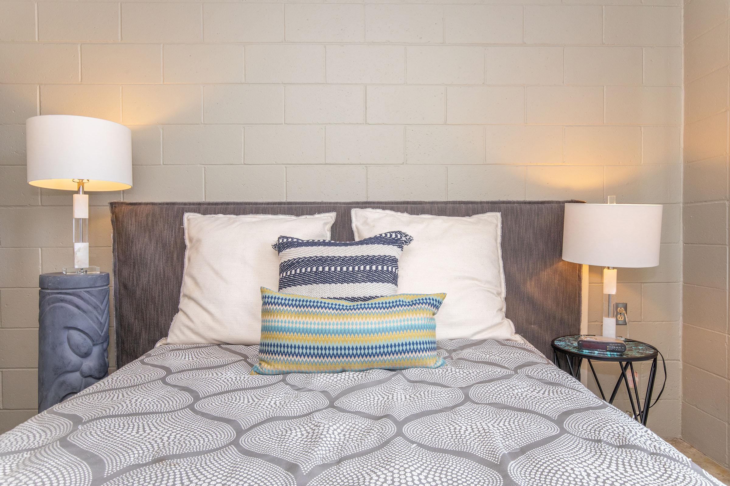 A cozy bedroom featuring a bed with decorative pillows and a gray patterned blanket. There are two bedside lamps providing warm light, with a small side table next to the bed. The walls are painted in a light color, creating a calm and inviting atmosphere.
