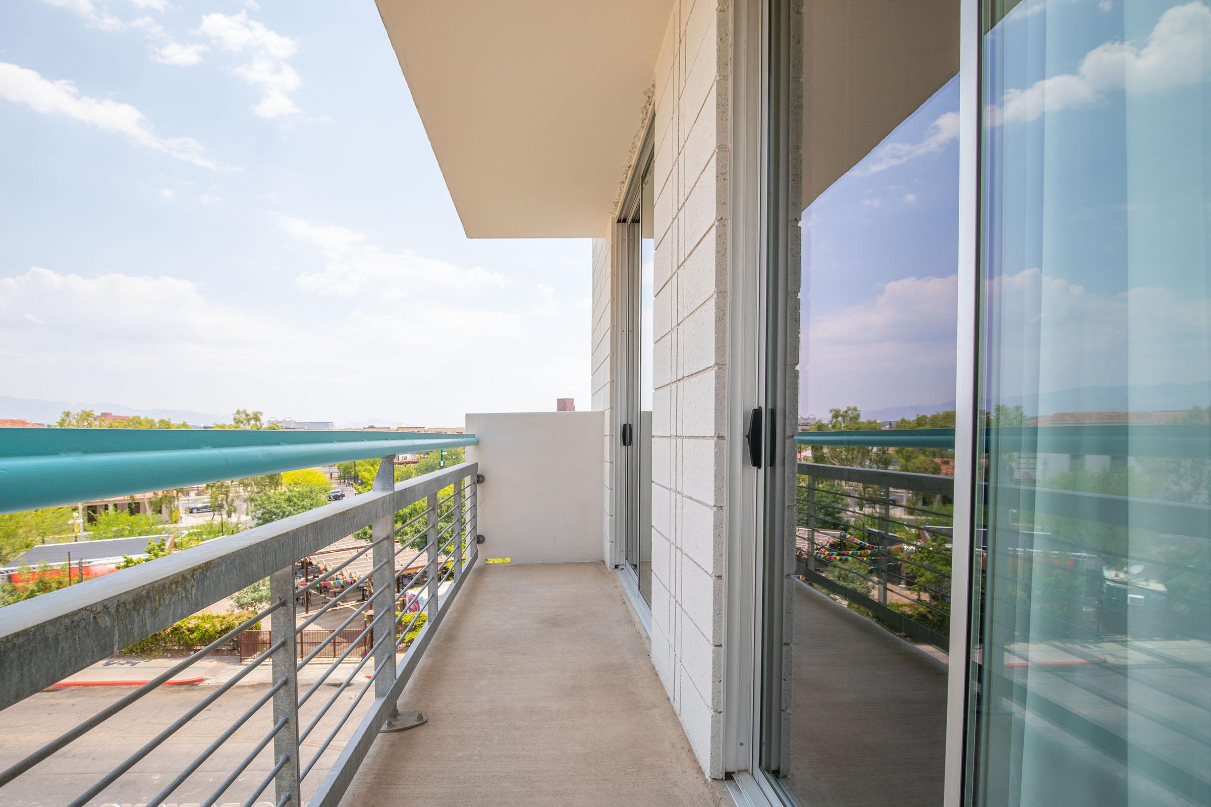 A clean, modern balcony with a railing, offering a view of an outdoor area. The balcony has a minimalist design, featuring glass doors and a concrete floor. Sunlight illuminates the space, highlighting the clear blue sky and some greenery in the background.