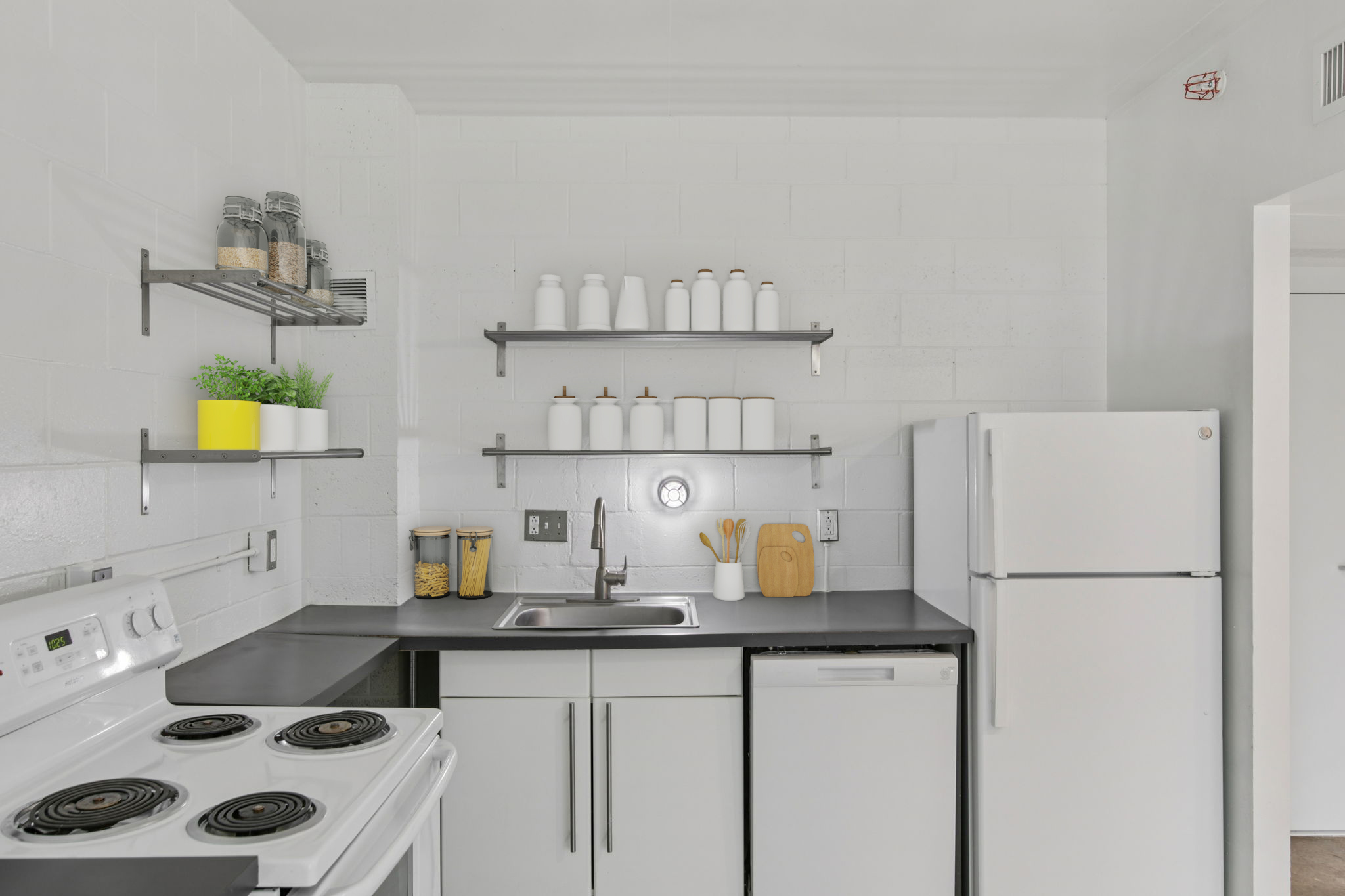 A modern kitchen featuring a white stove, refrigerator, and sink. There are open shelves with jars, white containers, and a small green plant. The walls are painted white, and the countertops are dark. Bright lighting enhances the clean, minimalist design.