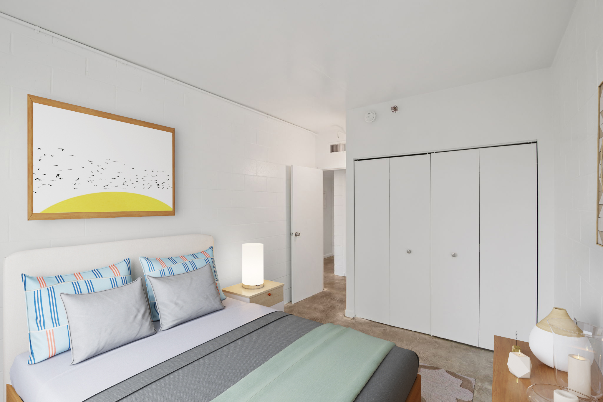 A bright, modern bedroom featuring a bed with striped pillows and a gray blanket. A minimalist artwork with birds is displayed on the wall. A wooden nightstand with a lamp is beside the bed, and sliding closet doors are visible in the background, along with a door leading to another room. The room has neutral-colored walls and carpet.