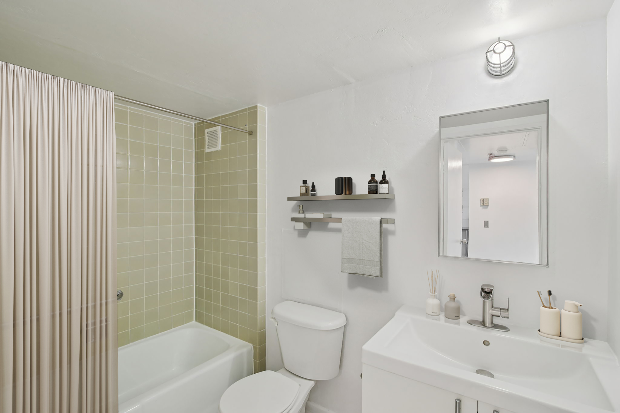 A modern bathroom featuring a light green tile shower area, white bathtub, and a sleek vanity with a sink. The space is well-lit with a ceiling fixture and includes a mirror above the vanity. Shelves display various toiletries, and a shower curtain is partially visible.