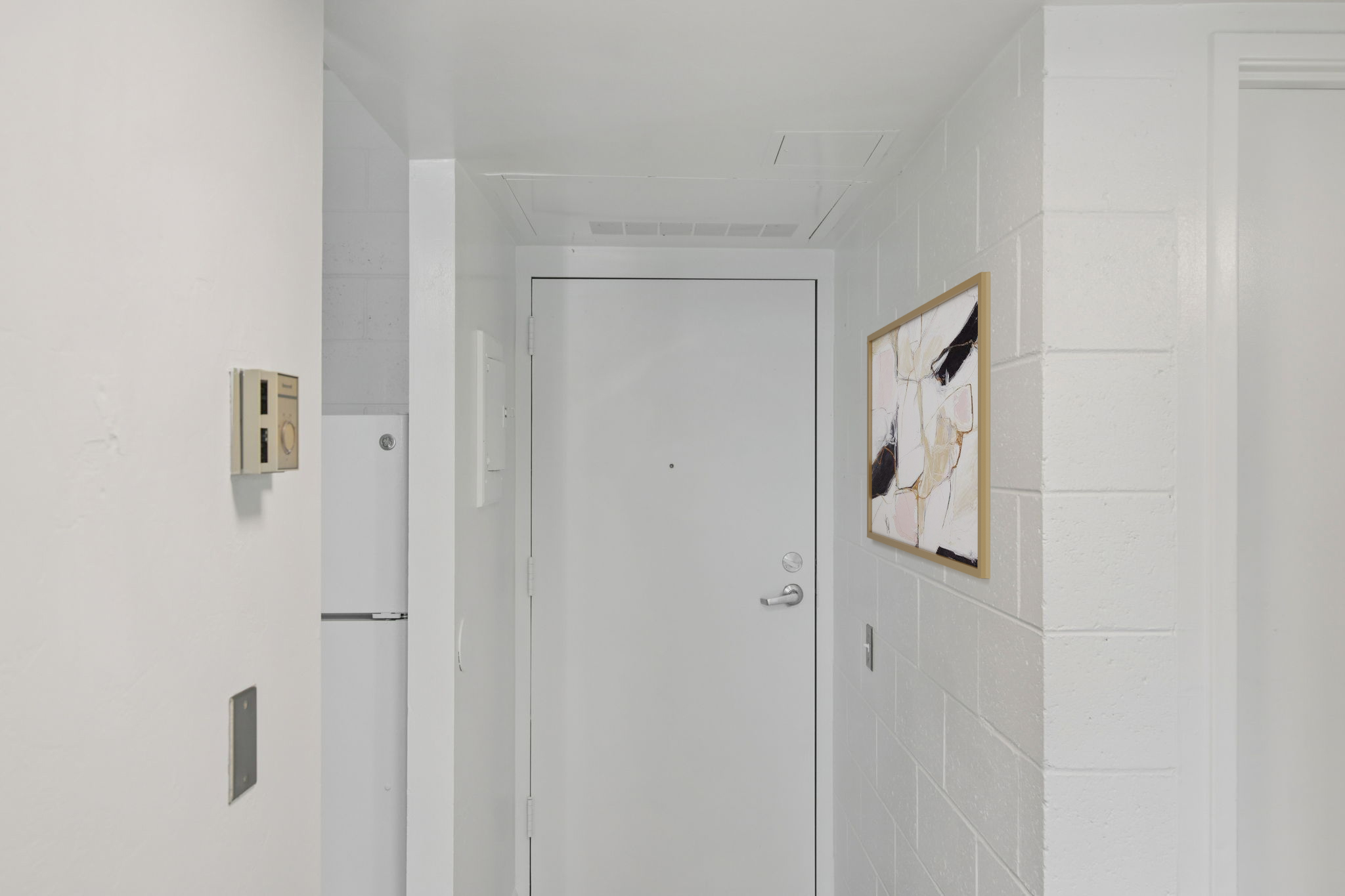 A clean, modern hallway with white walls and a closed door at the end. To the right, there is a framed abstract artwork. A refrigerator is visible in the background, adding to the minimalist aesthetic of the space.