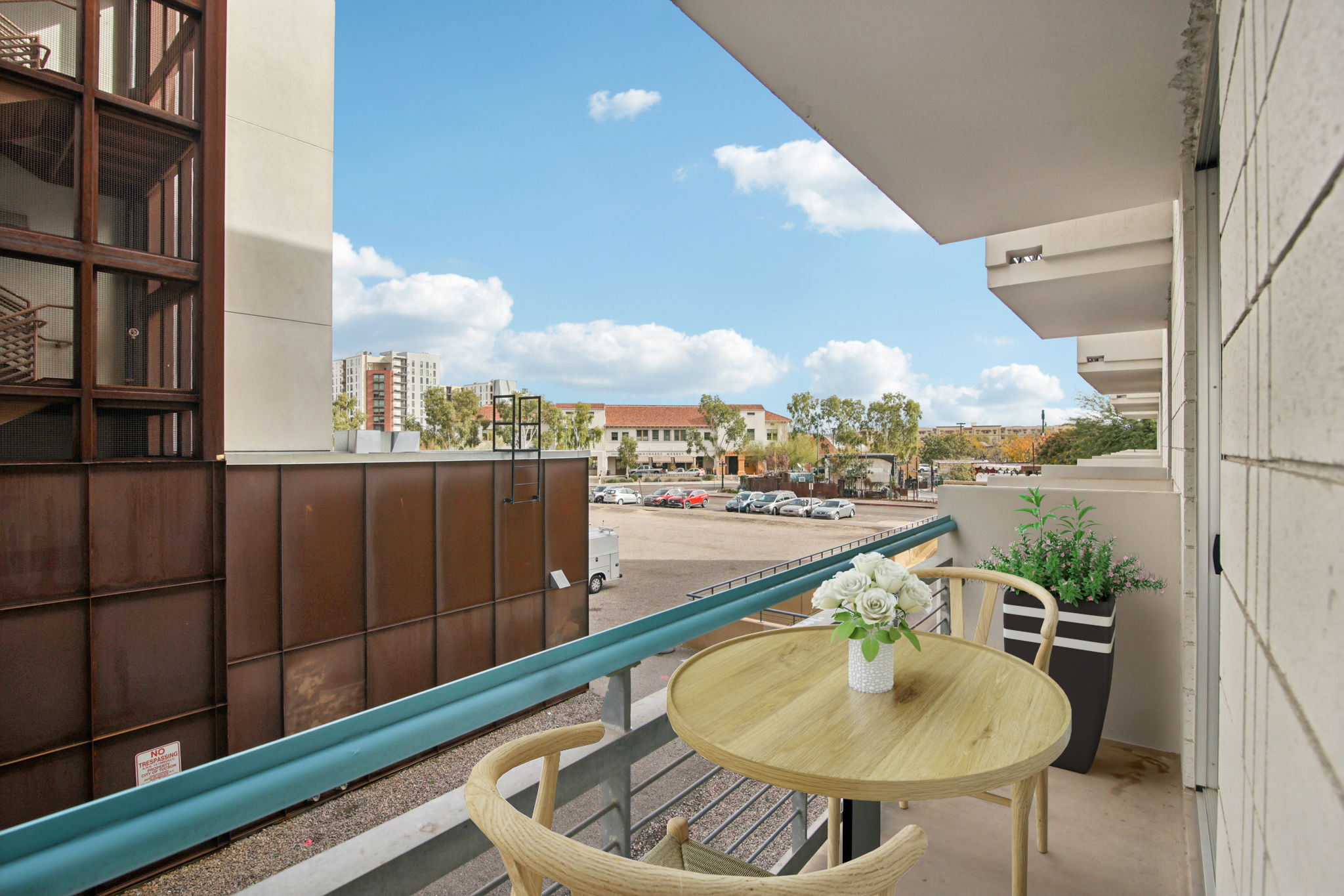 A balcony with a small round table and two chairs, adorned with a vase of flowers. In the background, a view of a parking lot and buildings under a clear blue sky with scattered clouds. The adjacent structures feature modern architecture.