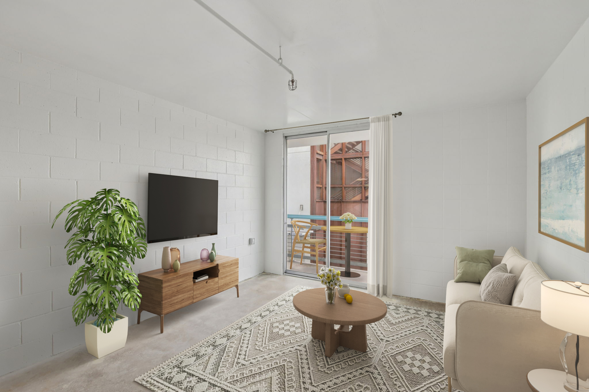 A modern living room featuring a beige sofa, a round wooden coffee table with decorative items, and a large flat-screen TV mounted on a wall unit. A potted plant adds greenery, while a sliding glass door leads to a balcony with outdoor seating. The space has light-colored walls and a patterned area rug.