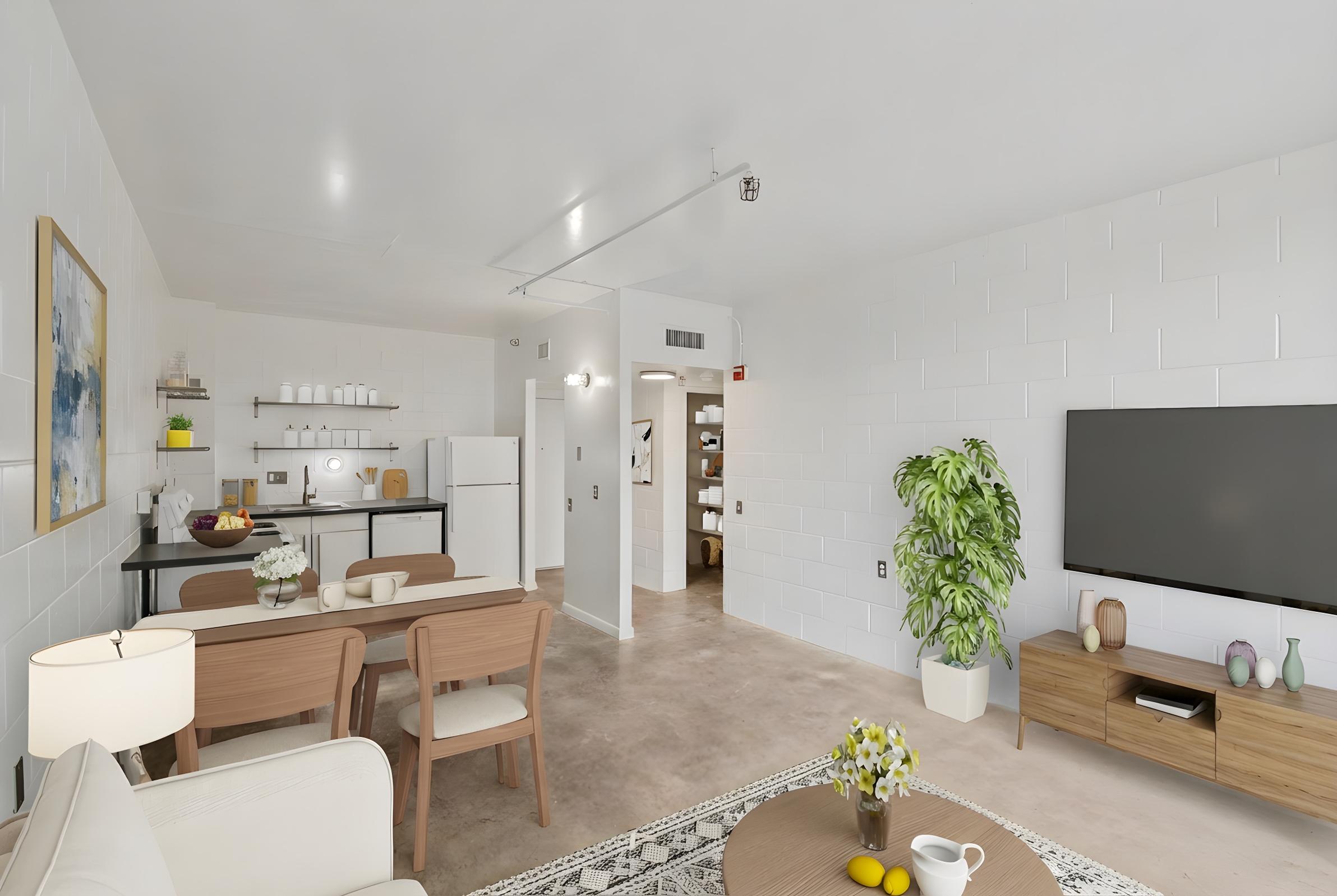 A modern, bright living space featuring a cozy seating area with a light sofa, a coffee table with decorative items, a dining table set for four, and a well-equipped kitchen. A large TV is mounted on the wall, and a potted plant adds a touch of greenery to the room. The walls are white with a minimalist design.