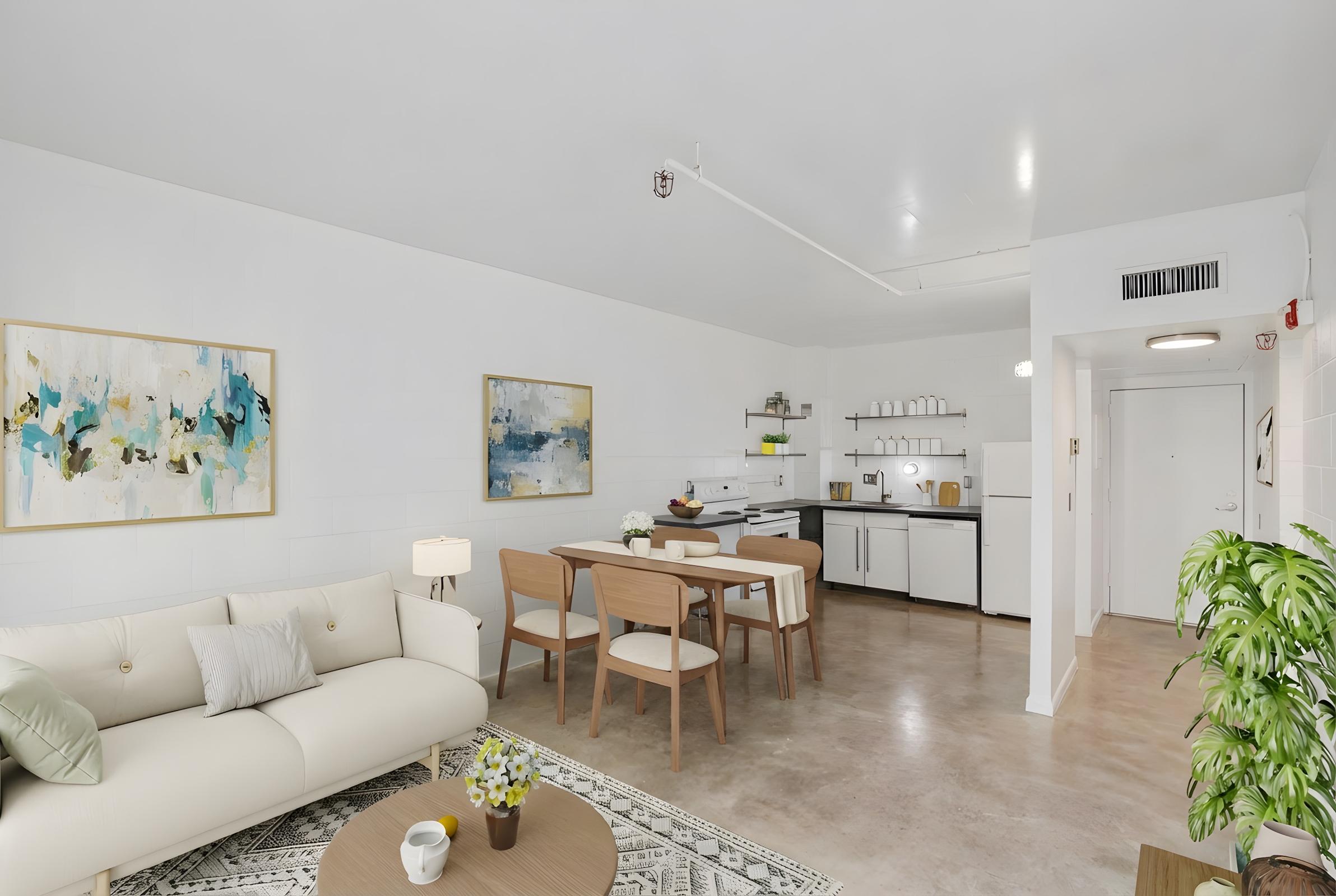 A bright, modern open-concept living space featuring a beige sofa, wooden dining table with chairs, and a minimalist kitchen. The walls showcase abstract art, and there's a decorative plant and small coffee table. The flooring is light-colored concrete, contributing to a clean, airy atmosphere.