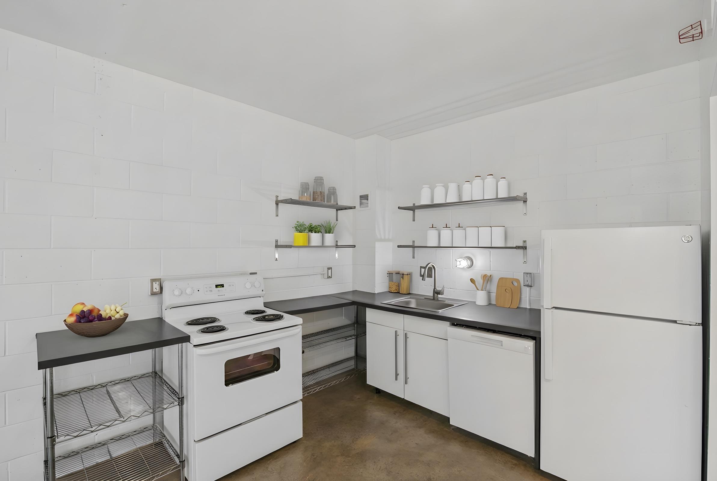 Modern kitchen featuring white cabinets and appliances, a black countertop, and shelves with jars. Includes a stove, sink, and a refrigerator. A fruit bowl is placed on a shelf, with a simple and clean design emphasizing functionality and minimalism.
