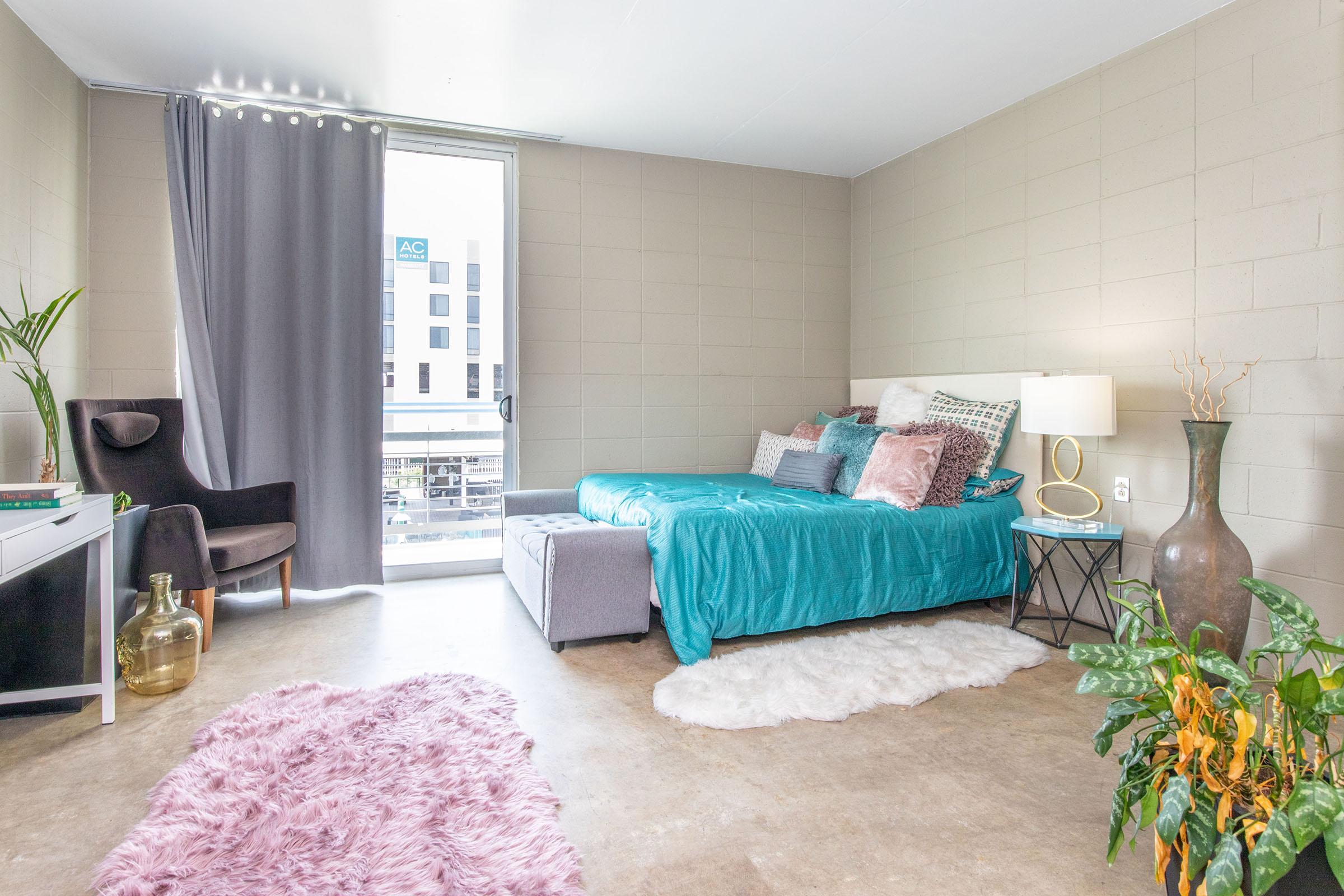 A cozy, modern bedroom featuring a turquoise bedspread, decorative pillows, and a gray throw blanket. There's a large window with curtains, a stylish armchair, a small side table with a lamp, a potted plant, and a fluffy pink rug on the floor, creating a warm, inviting atmosphere.
