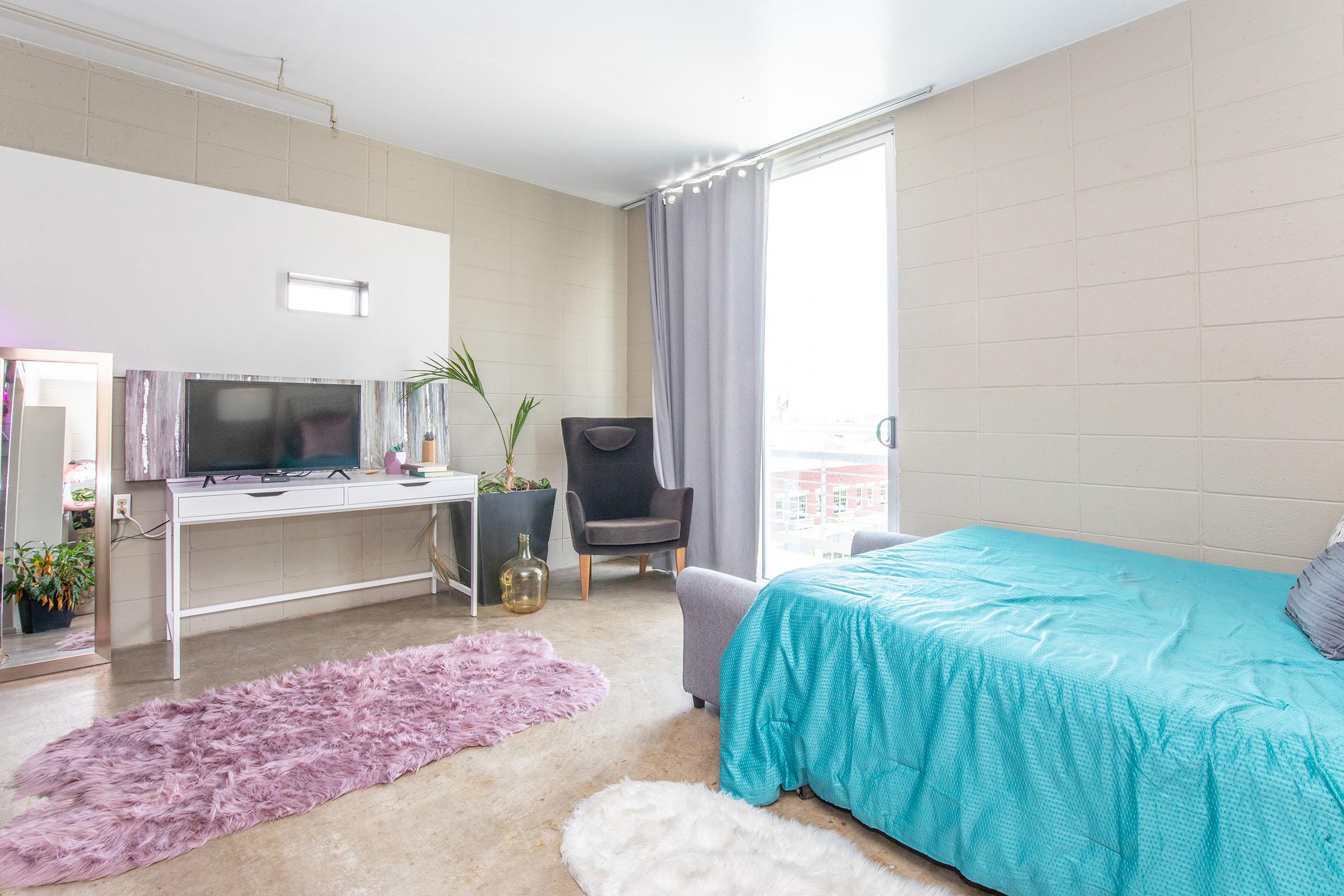 A bright, minimalist room featuring a bed with a turquoise cover, a soft purple rug, a black armchair, and a white desk with a TV. Natural light filters through a large window, complemented by a green plant and decorative elements in the space, creating a cozy and inviting atmosphere.