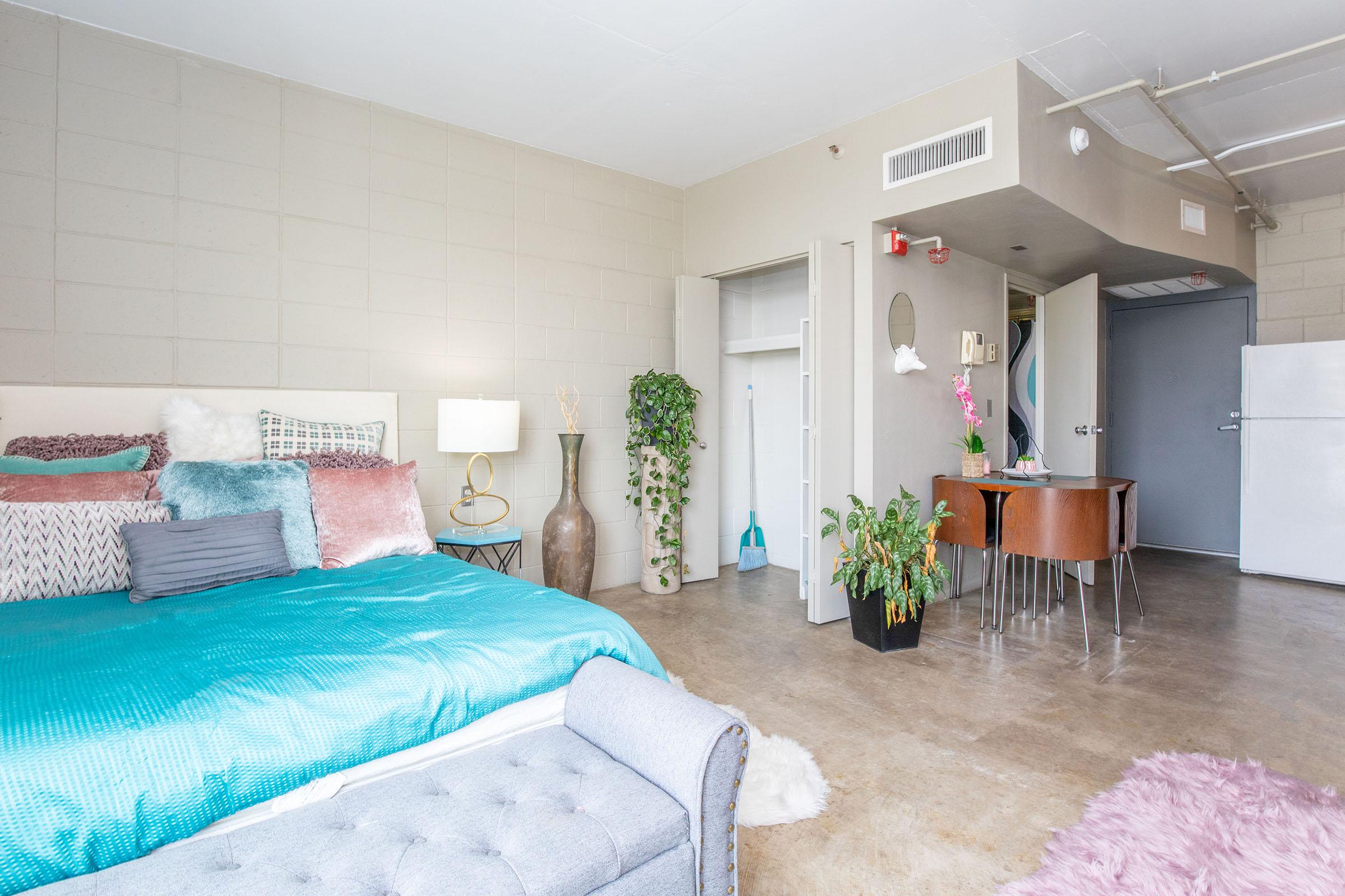 A brightly lit studio apartment featuring a bed with colorful bedding, a decorative plant, a small dining set, and a white refrigerator. The walls are a neutral tone, and there's a closet space visible with a few hangers. The overall decor gives a cozy and modern vibe.