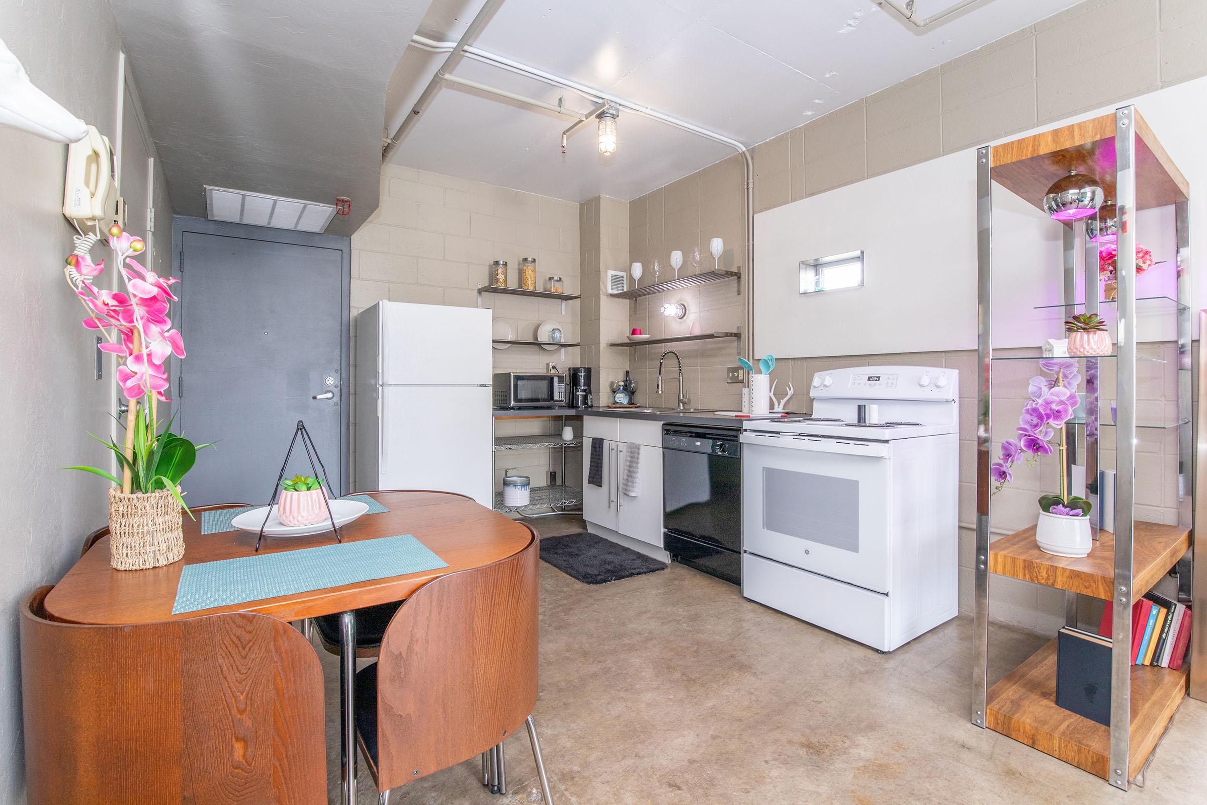 A modern kitchen with a white stove, refrigerator, and microwave. There is a wooden dining table set for two, featuring a teal placemat and decorative plants. Shelves display potted orchids. The walls are light-colored, and the flooring is concrete, giving a contemporary feel to the space.
