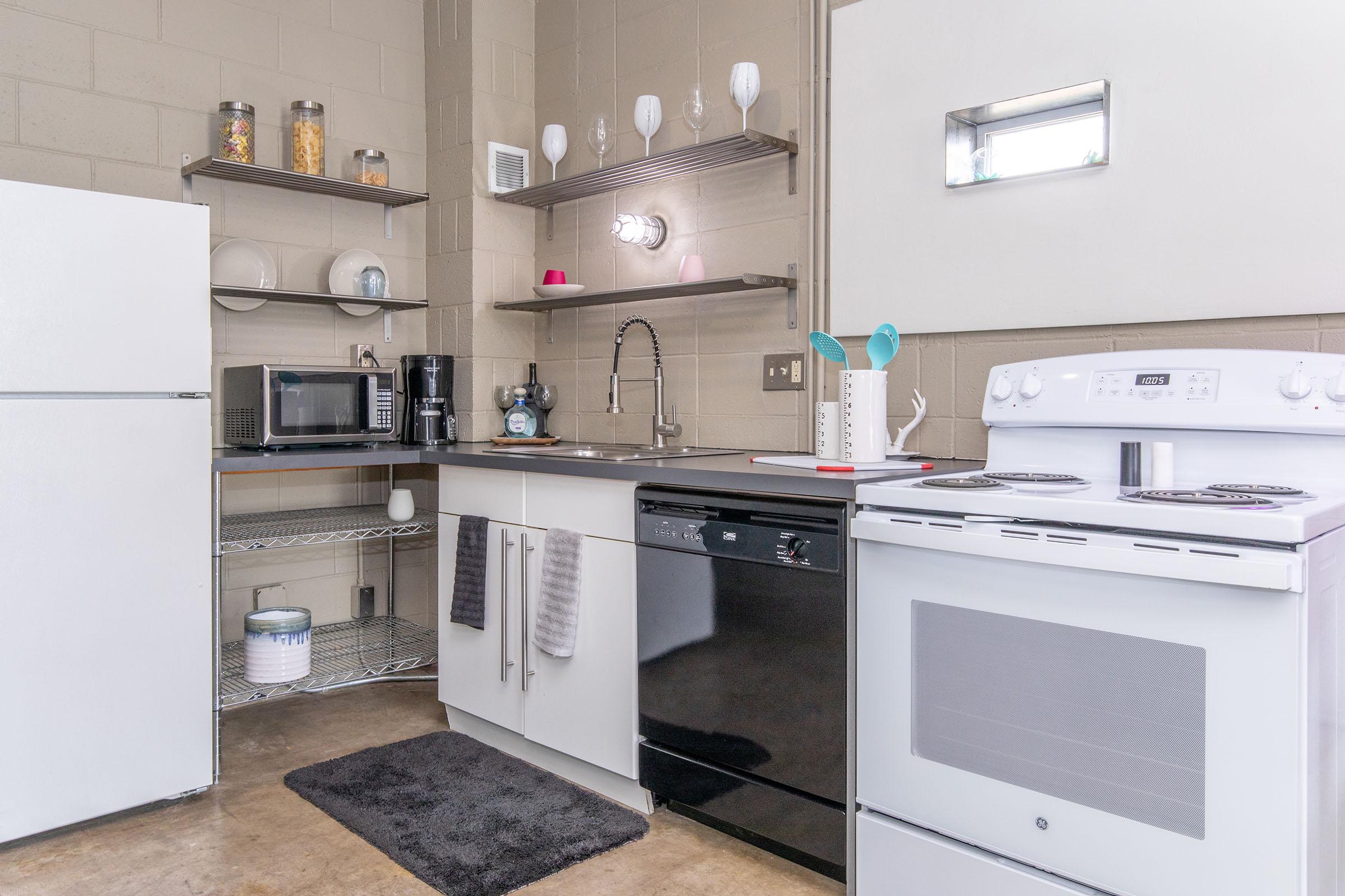 A modern kitchen featuring a white refrigerator, a stainless steel dishwasher, an electric stove, and a microwave. Shelves above the countertop hold dishes and glasses. A coffee maker is next to the sink, and a black mat is placed on the floor in front of the sink. The walls are painted a muted color.
