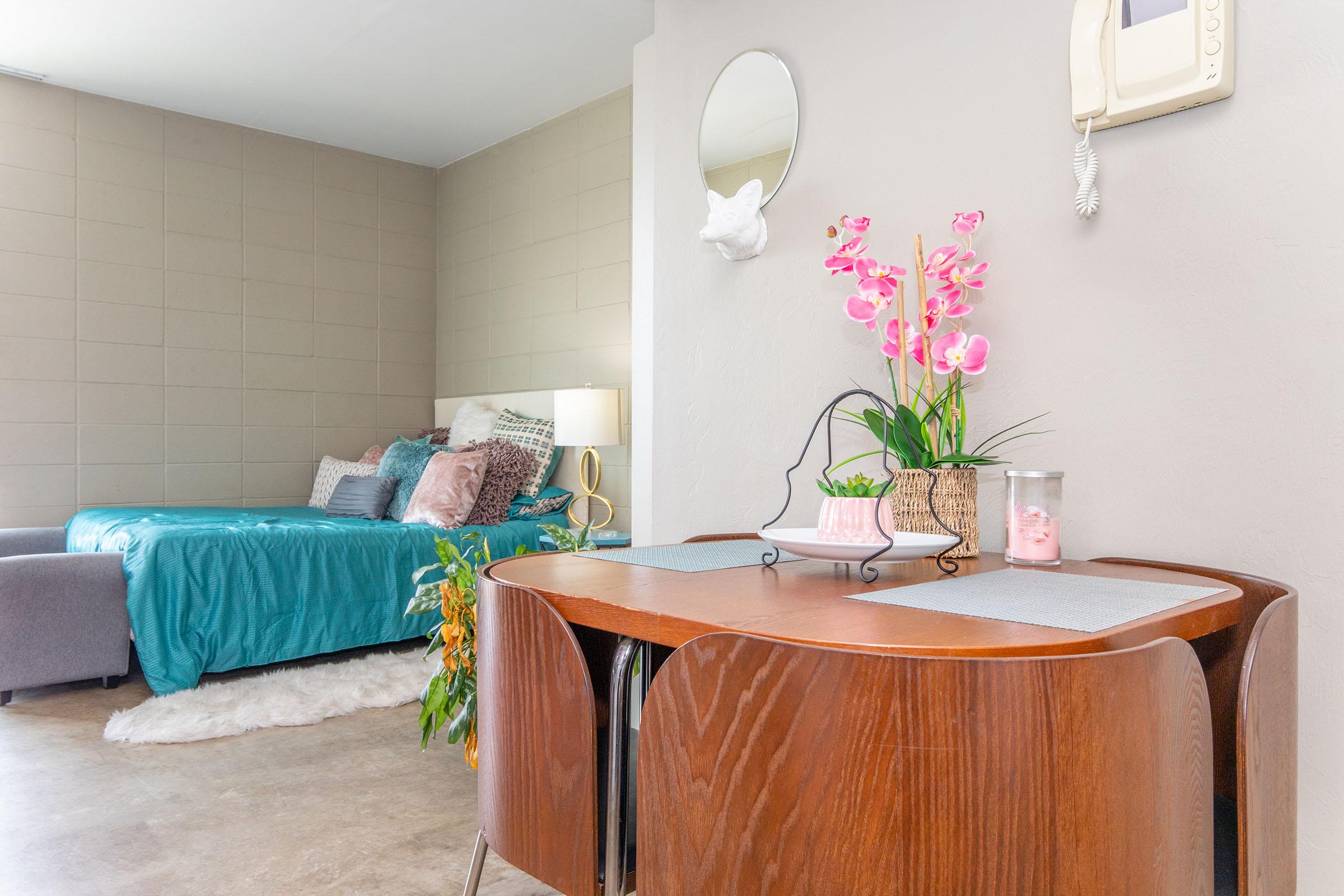 Cozy interior of a small studio apartment featuring a turquoise bedspread, decorative pillows, a small dining table with a floral centerpiece, and a mirror on the wall. The room has modern decor with soft lighting and a plush rug, creating a welcoming atmosphere. A vintage-style phone is mounted on the wall.