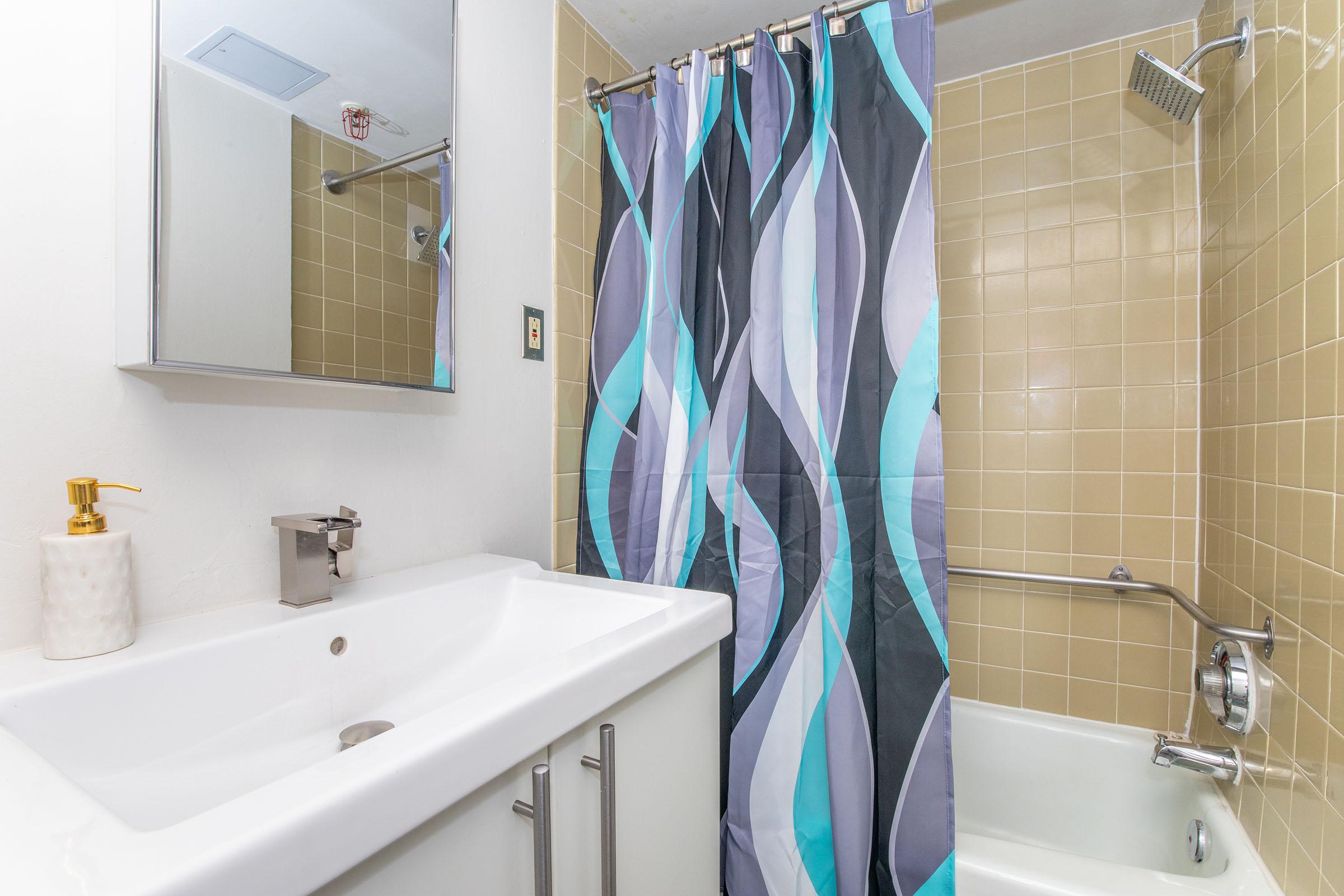 A small bathroom featuring a modern sink with a sleek faucet, a mirror above, and a shower area with a colorful wavy shower curtain. The walls are tiled in a light beige color, and a bath tub is visible beside the shower. There is a soap dispenser on the counter, adding to the clean, organized appearance.