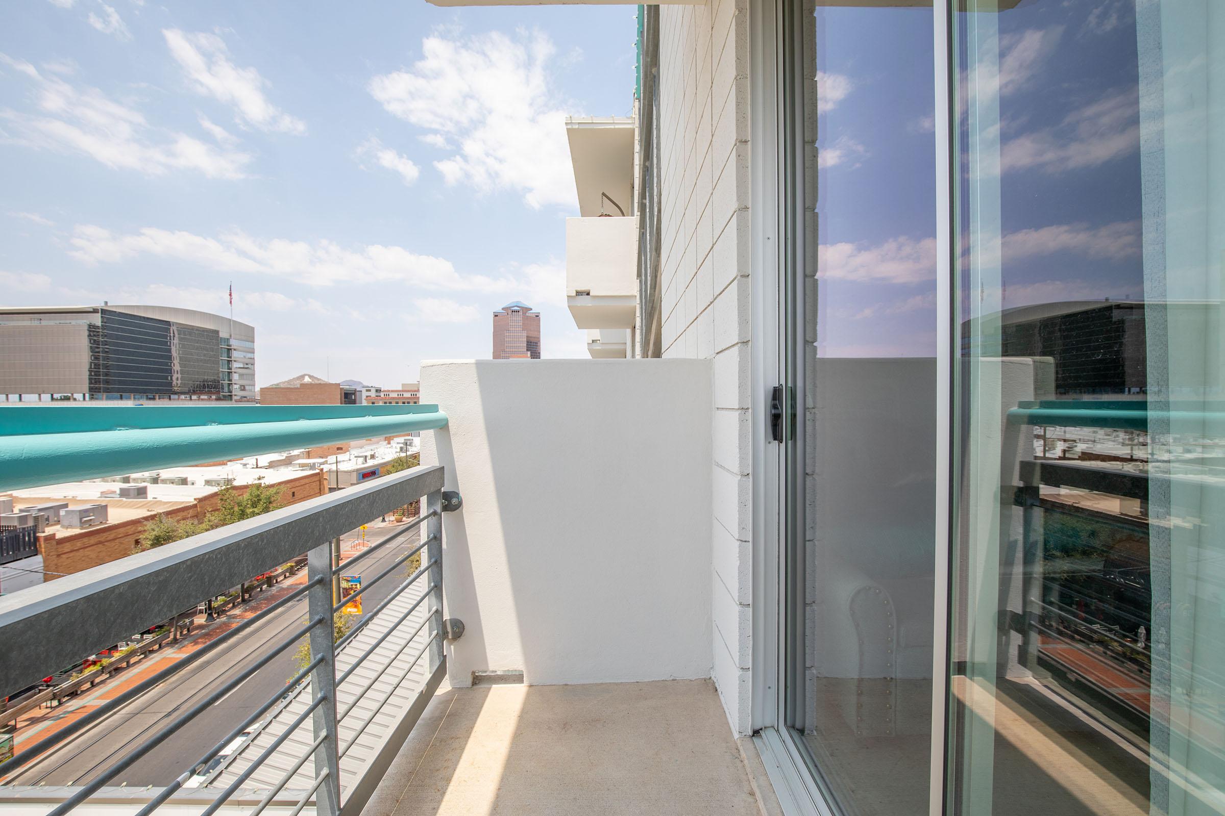 View of a balcony with a glass railing, overlooking a cityscape with modern buildings. A clear sky with some clouds is visible. The balcony is clean and minimally furnished, providing a spacious feel. The scene suggests an urban environment with bustling streets below.