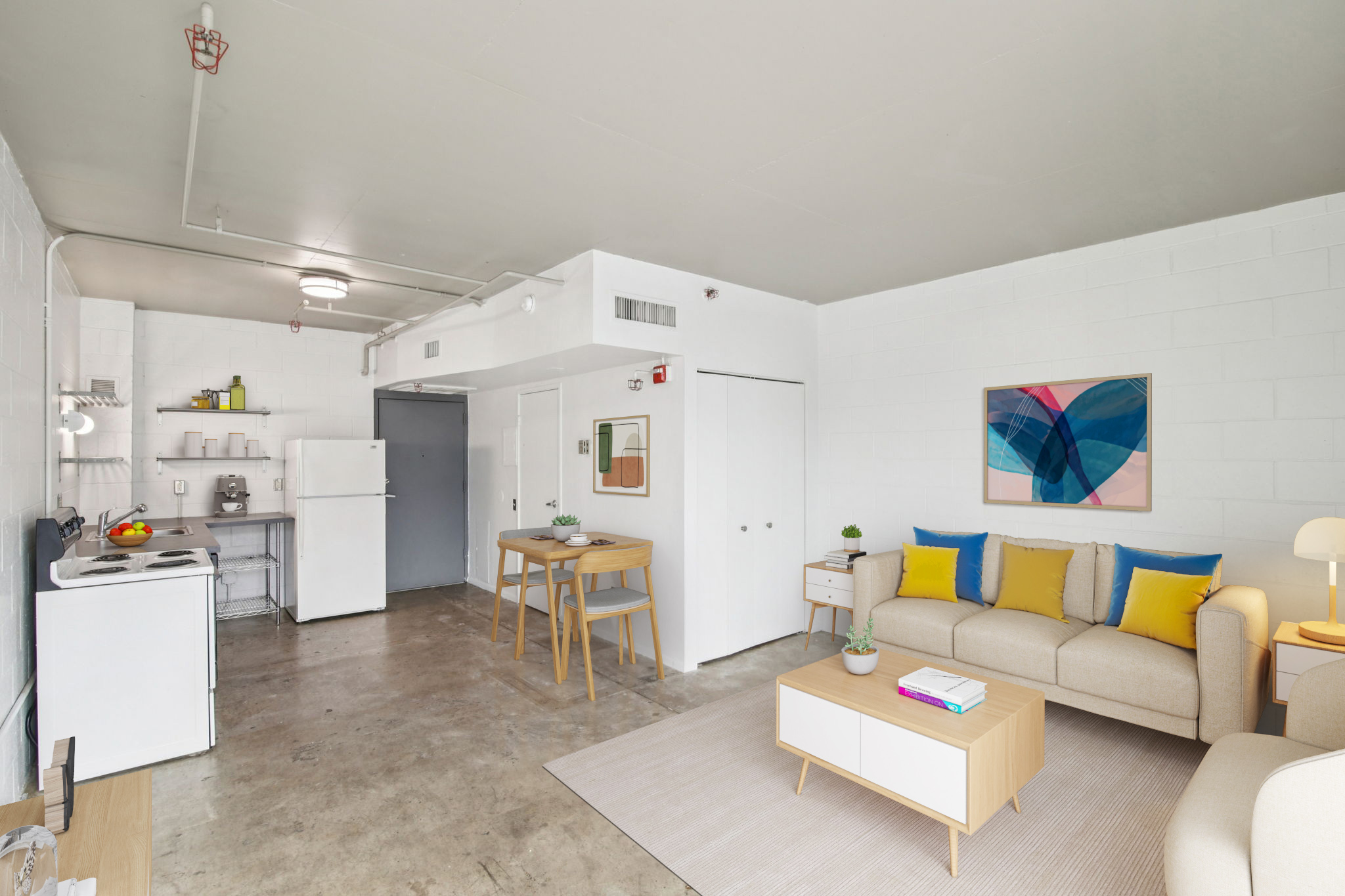 A modern, minimalist living space featuring a cozy sofa with yellow cushions, a light wood coffee table, and a dining area with a wooden table and chairs. The kitchen area includes a refrigerator and shelves, with a colorful abstract artwork on the wall. The floor is concrete, and natural light fills the room.