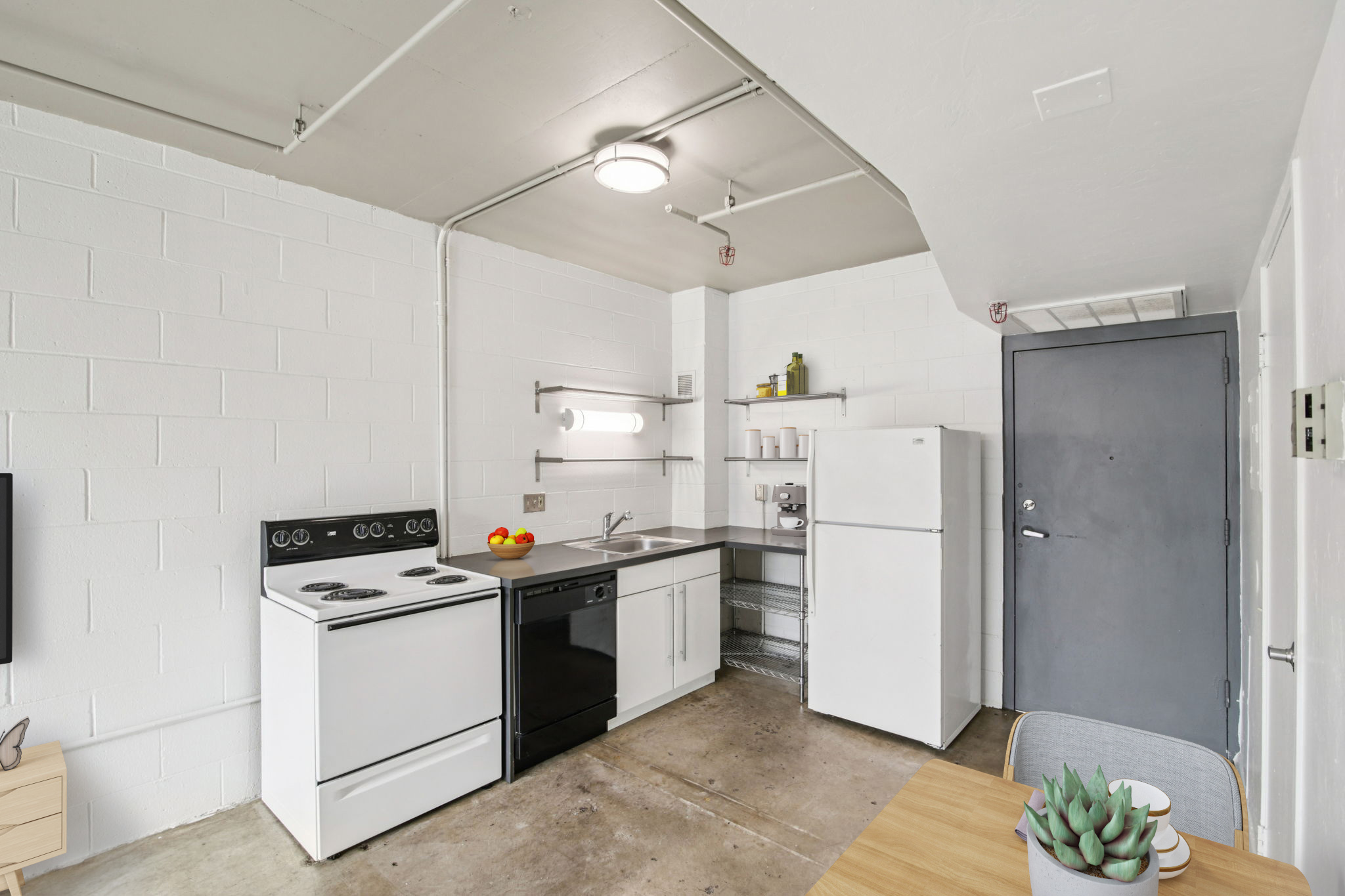 A compact kitchen featuring a white stove and oven, a black dishwasher, and a white refrigerator. The kitchen has a minimalist design, with a countertop, basic shelving, and light-colored walls. A wooden dining table is visible in the foreground, adding a touch of warmth to the space.