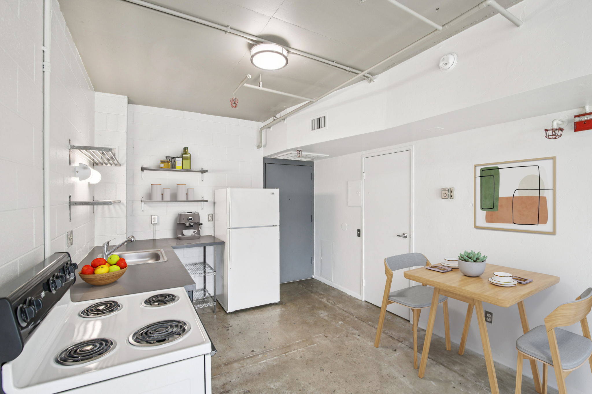 A modern kitchen featuring a white refrigerator, stove, and sink. The walls are painted white, and there's a small dining table with two chairs. A bowl of colorful fruit is on the table, and decorative art hangs on the wall. The floor has a concrete finish, creating an industrial feel.