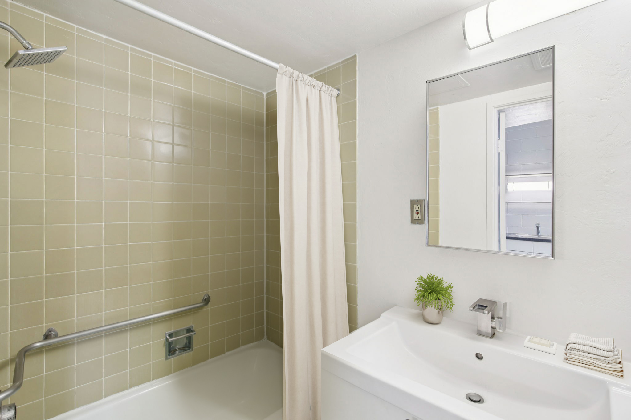 A clean bathroom featuring a bathtub with a showerhead and curtain, light green tiled walls, a modern vanity with a sink, and a mirror. Decorative green plant and toiletries are neatly arranged on the countertop. A towel is placed beside the sink, enhancing the fresh and inviting atmosphere.