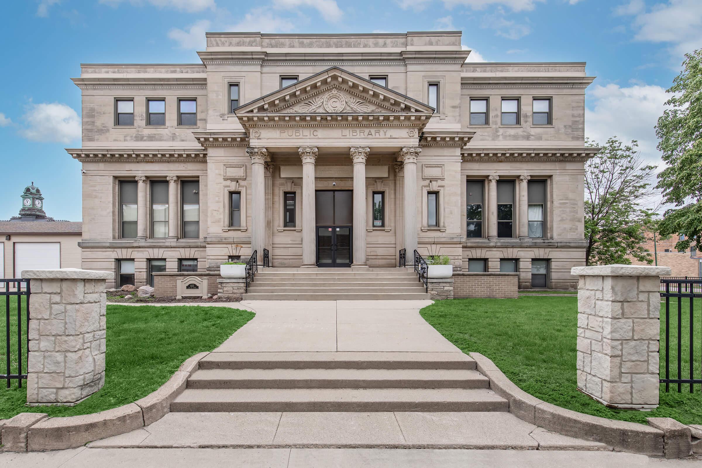 An elegant stone building with a grand facade featuring columns and intricate architectural details, identified as a public library. A wide concrete pathway leads up to the entrance, flanked by well-maintained grass and shrubs. The sky above is clear and blue, adding to the inviting atmosphere of the structure.