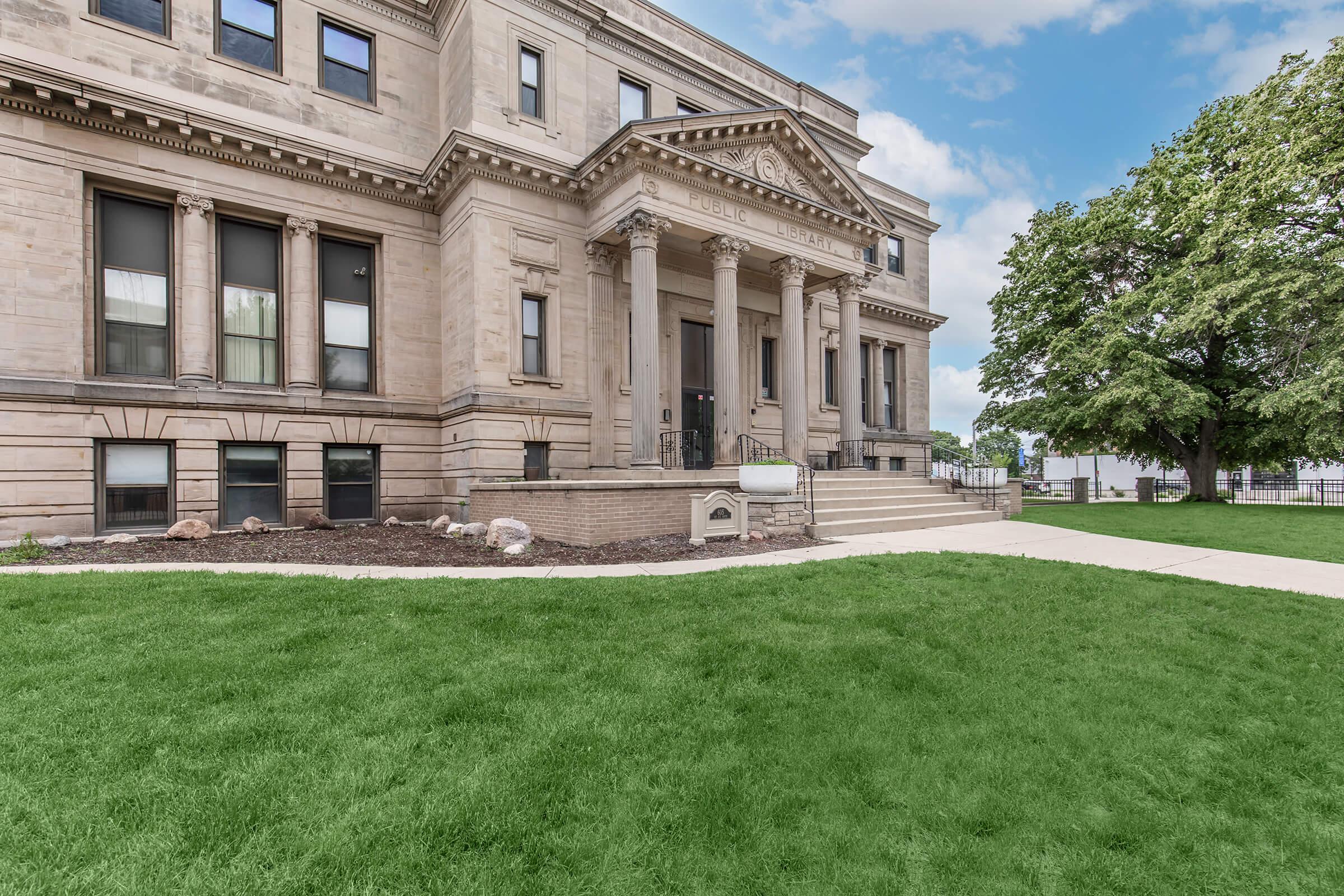 Historic building with classical architecture featuring decorative columns and intricate stonework. Surrounding it is a well-manicured green lawn with a pathway leading to the entrance. Trees are visible in the background under a blue sky with scattered clouds.