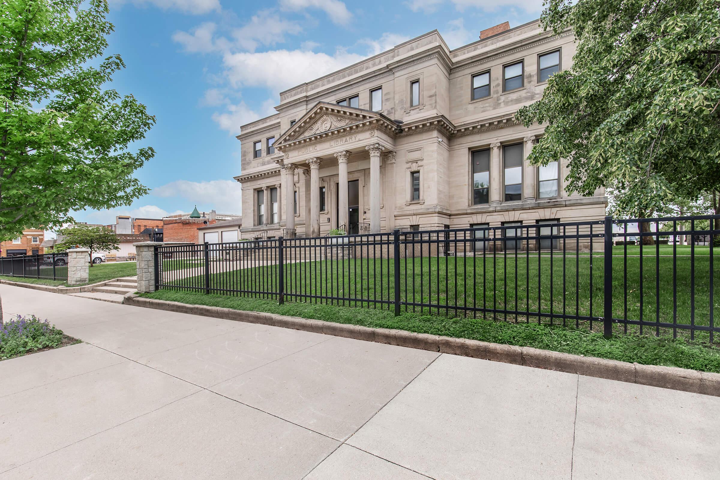 A grand building with a classical architectural design, featuring a large entrance with columns. The building is surrounded by a green lawn and a black metal fence. A paved walkway leads to the entrance, with trees and a clear blue sky in the background.
