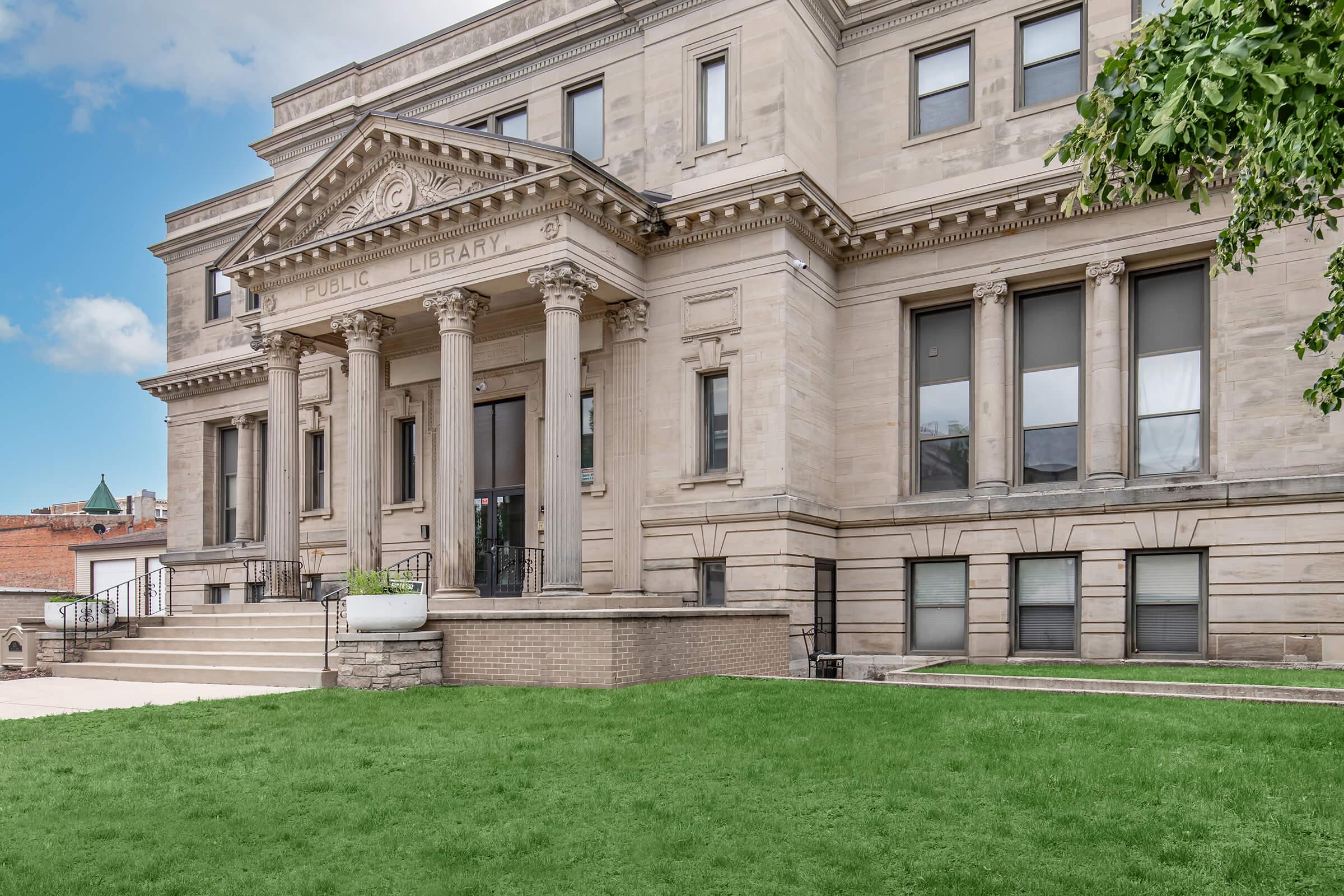 Facade of a stately public library featuring classical architectural elements like columns and large windows, set against a clear blue sky. The building is flanked by manicured green grass, creating a welcoming entrance with stone steps leading up to the main doors.
