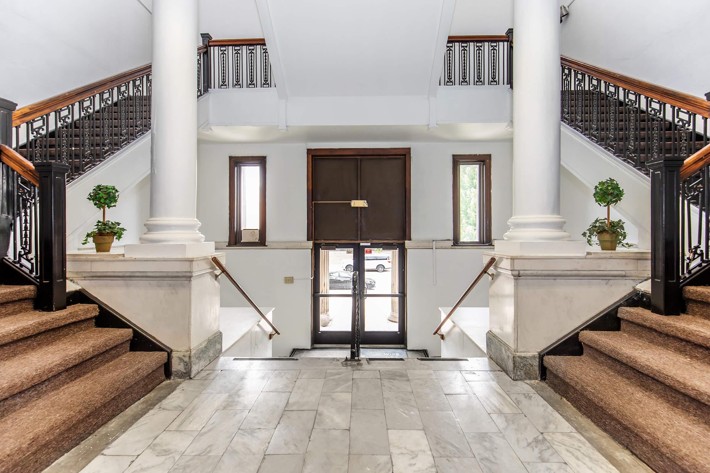 Interior view of a grand staircase in a building, featuring two sets of stairs leading to an entrance. The space is illuminated by natural light, with decorative columns and plants in pots on either side. A door is visible at the top of the stairs, providing access to the outside.