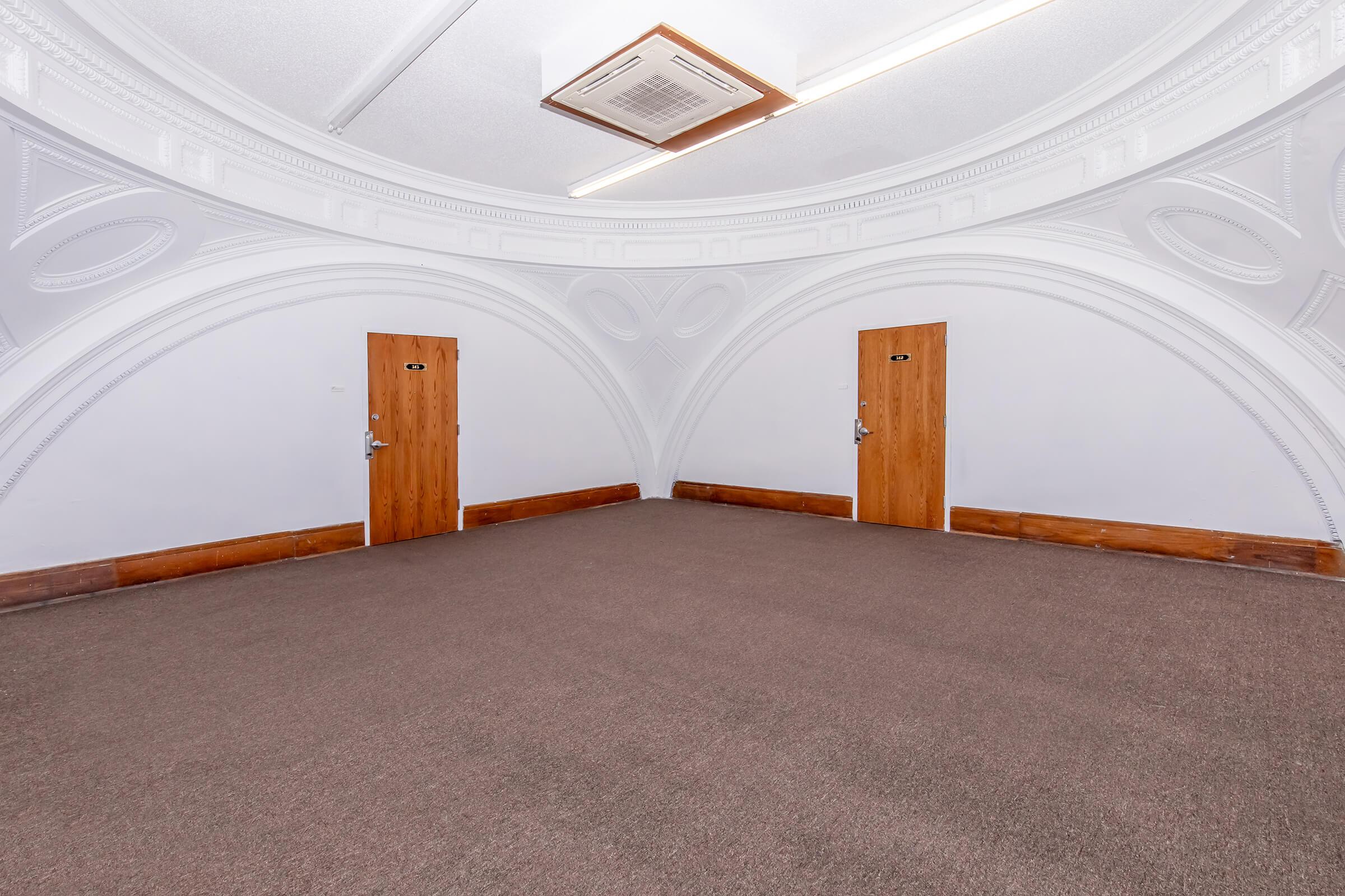 An empty room with a curved ceiling and a carpeted floor. There are two wooden doors on opposite walls, both featuring small signs. The walls are adorned with decorative moldings, and the overall ambiance is bright and spacious.