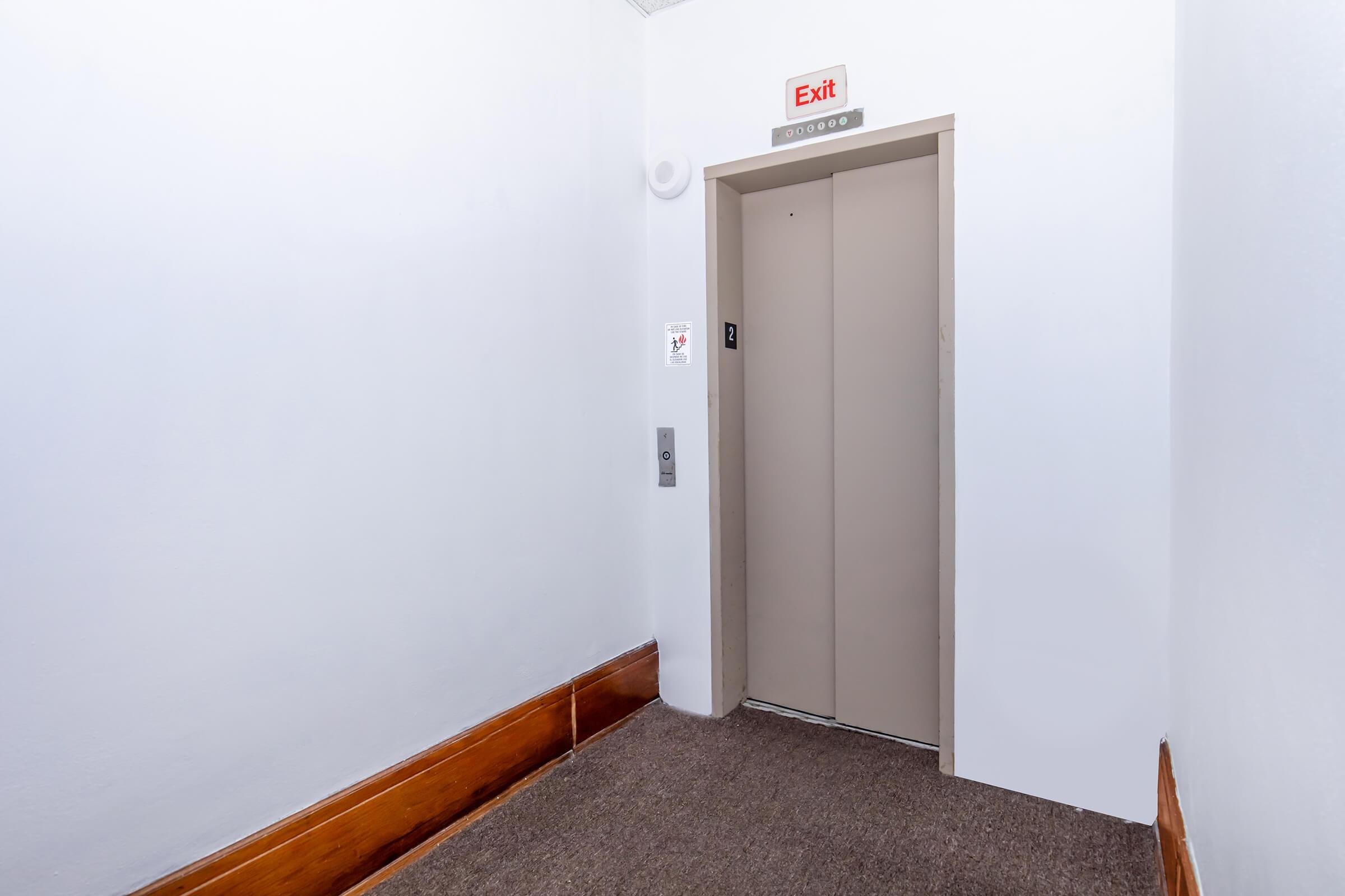 An empty elevator hallway featuring a closed elevator door. The wall is mostly white with a wooden panel at the bottom. There is an "Exit" sign above the elevator, and a control panel next to the door. The floor is covered with gray carpeting.