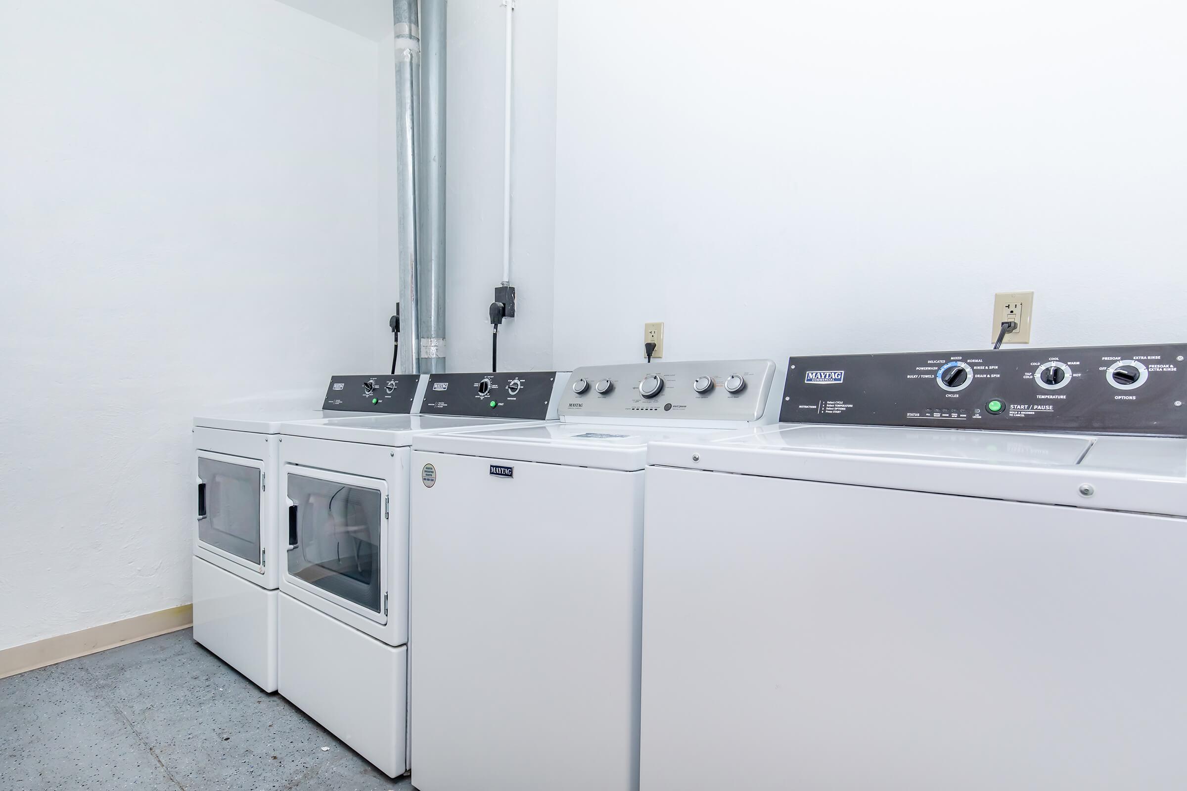 A clean laundry room featuring a stacked washer and dryer unit alongside a separate washing machine. The machines are white with control panels visible, and the walls are painted light colors. A vent pipe is visible on the left side, suggesting proper ventilation.