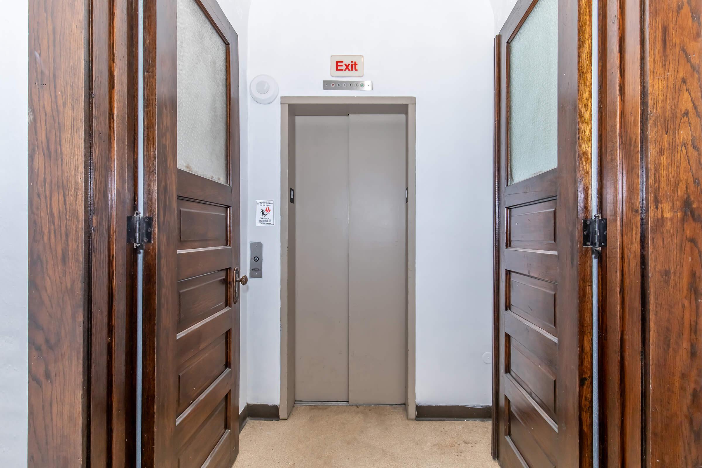 A hallway with two wooden swing doors leading to an elevator. The elevator doors are gray, and there is an exit sign above. The walls are white, and the floor is light beige carpet. The wooden doors have glass panels, and there are safety signs nearby.