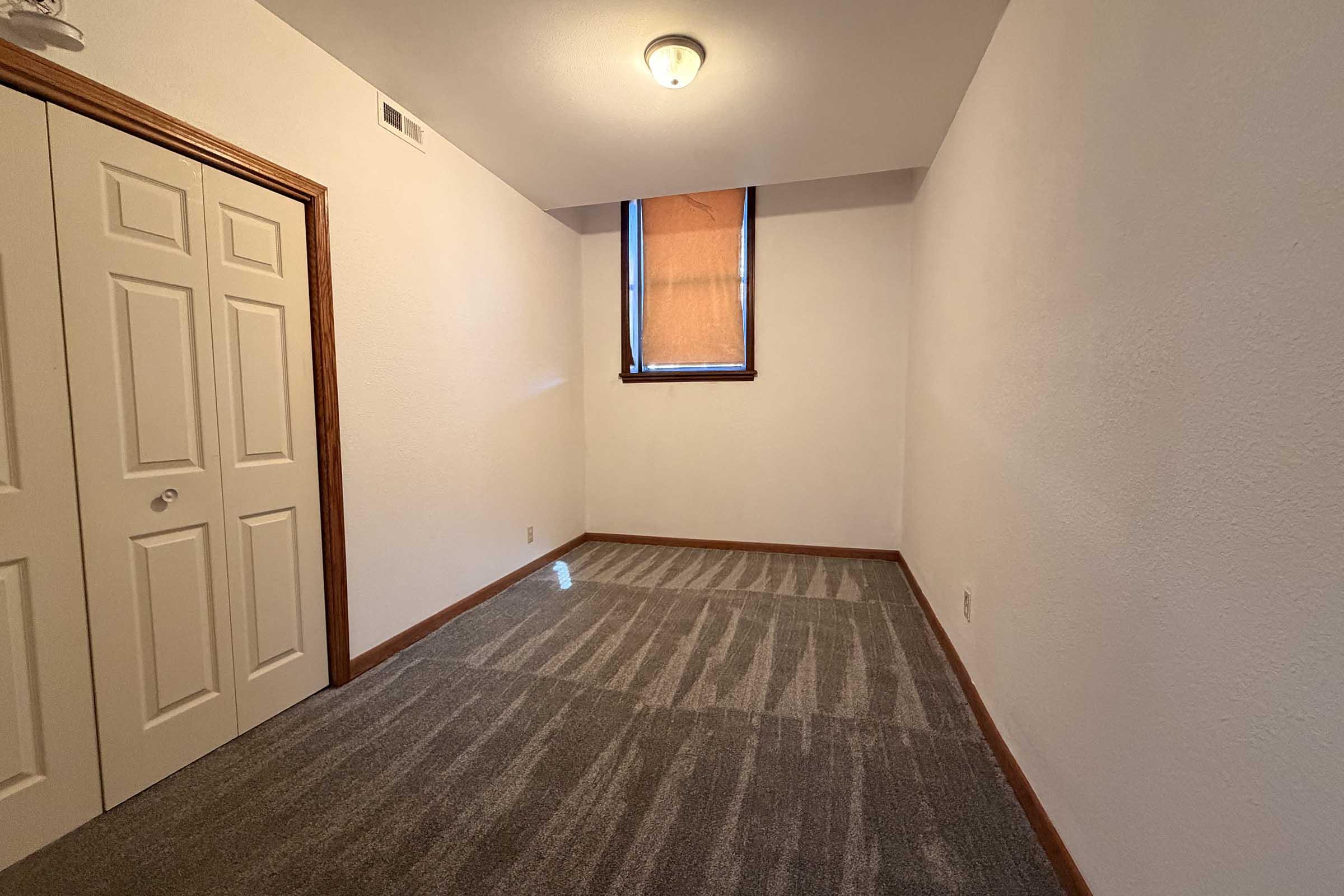 A small, empty room with light-colored walls and a window covered by an orange blind. The floor is carpeted in a muted gray pattern. There are double doors on one side, leading to a closet or storage area. The ceiling features a single light fixture, providing soft illumination.